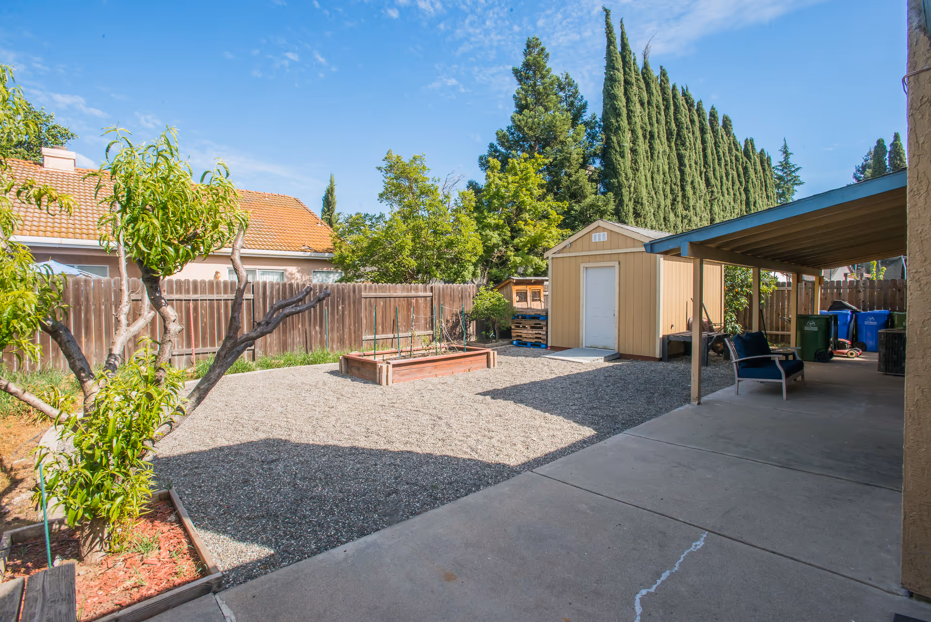 Backyard with gravel, trees, a storage shed, raised beds, and a shaded seating area.