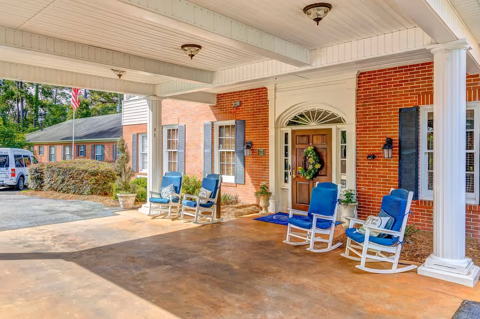 Covered entrance area of a brick building with four blue cushioned rocking chairs arranged near the front door, which is decorated with a green wreath. There are white columns supporting the roof of the entrance, potted plants near the door, and an American flag visible in the background along with a white van parked nearby.