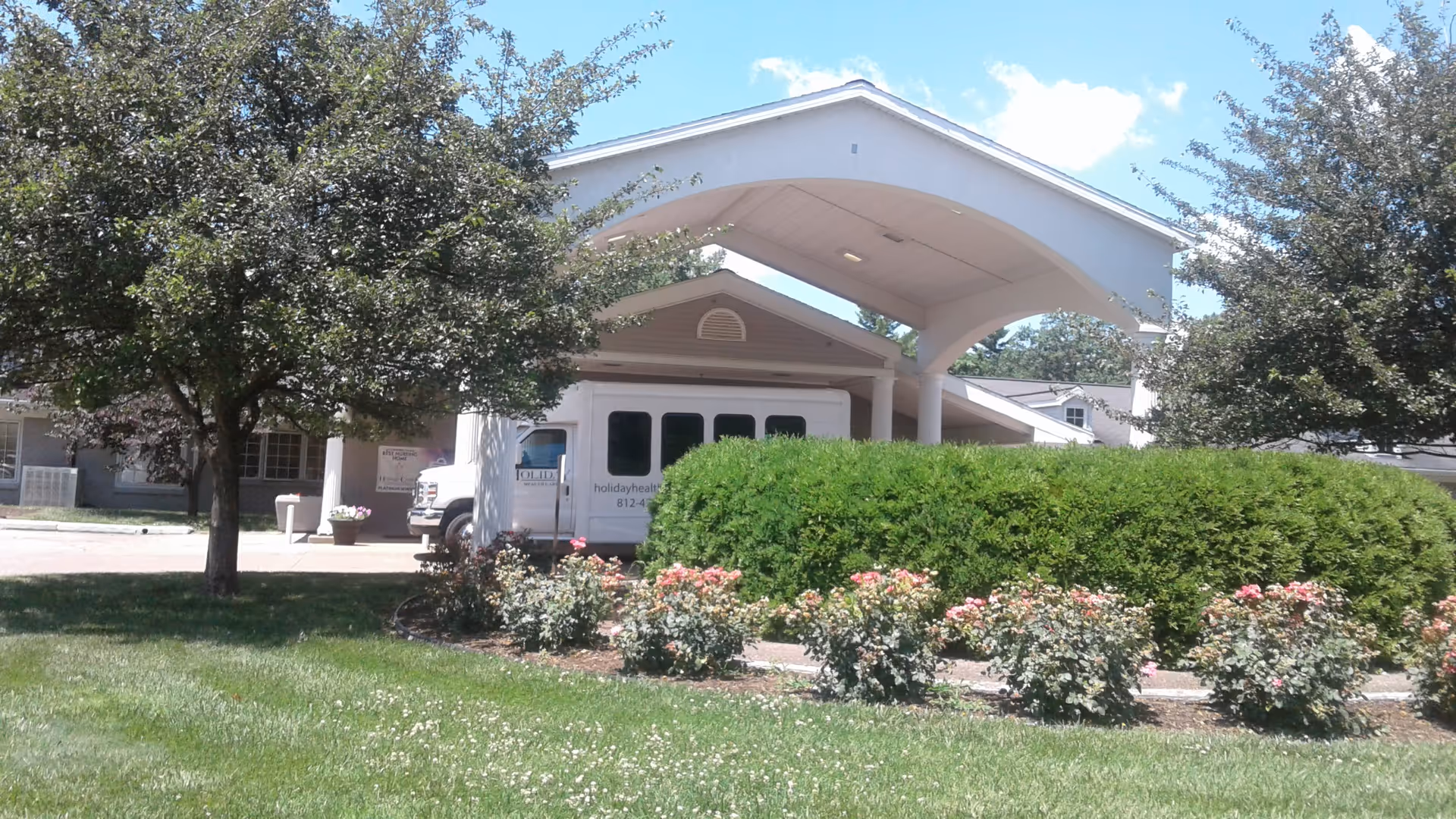 Exterior view of a senior living facility with curved entrance canopy, van, trees, and shrubs.