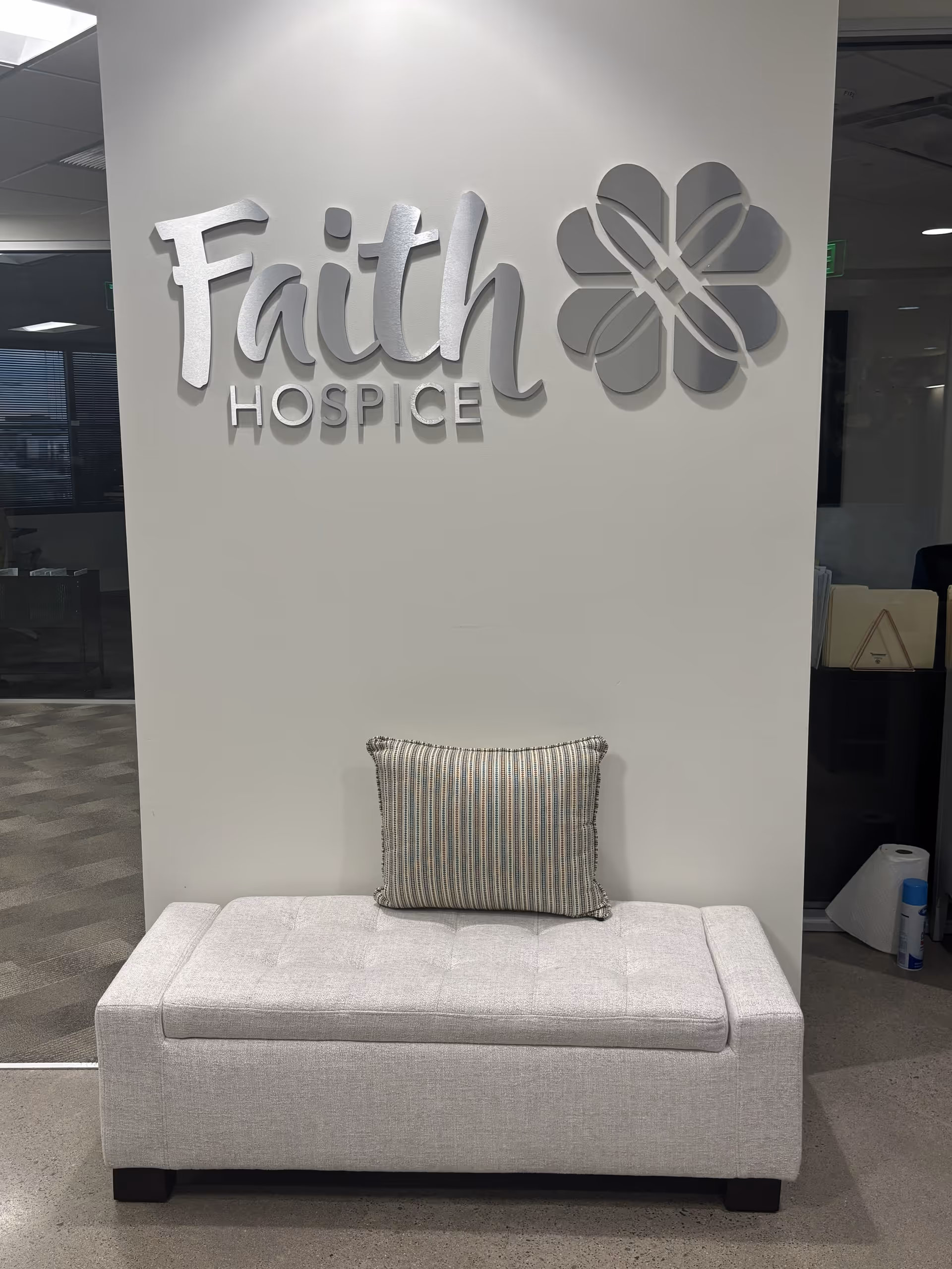 Light-gray bench in a lobby area beneath a Faith Hospice logo mural.