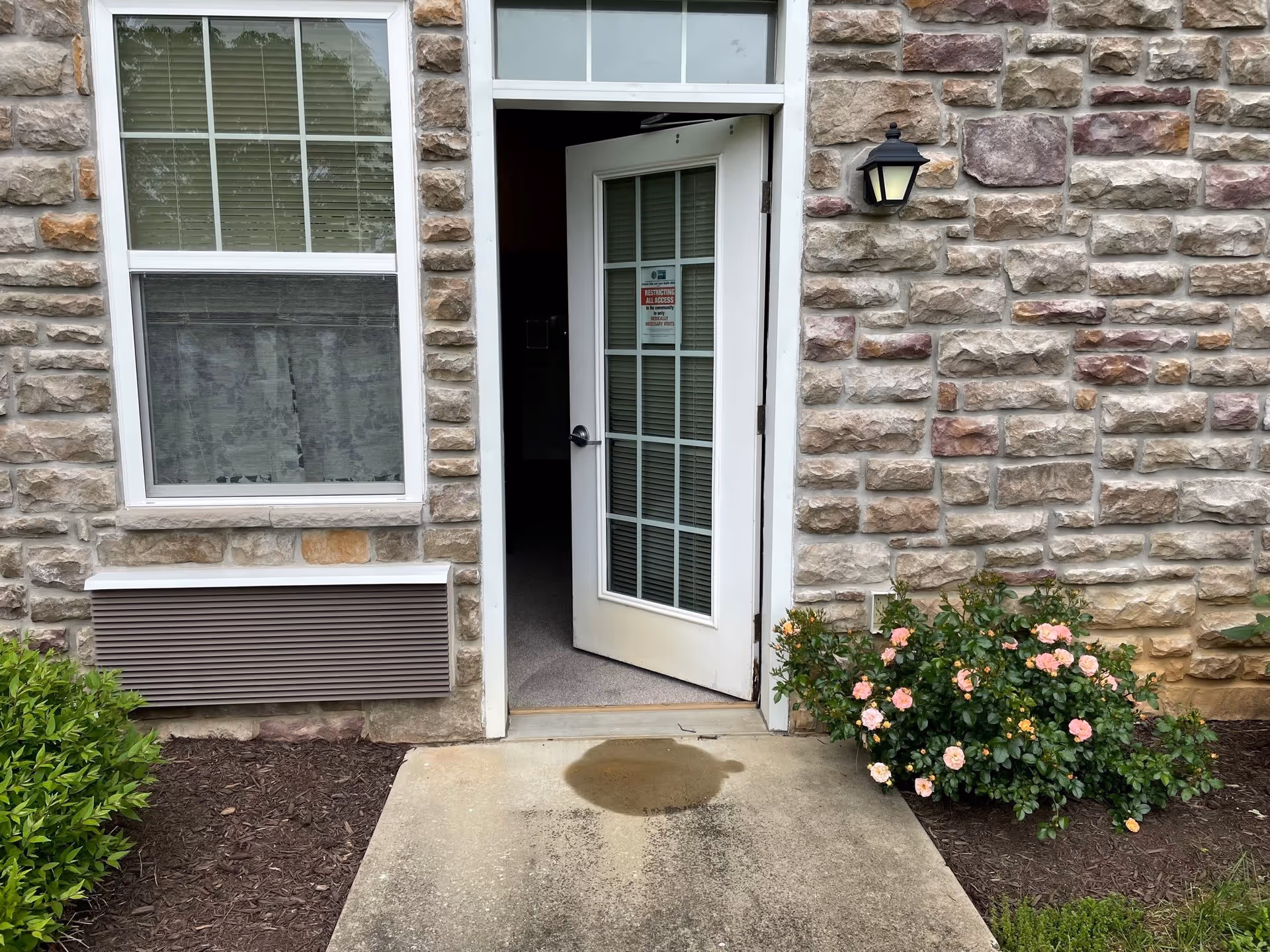 Stone-faced building entrance with a partially open glass-paned door, adjacent window, wall light, and a flowerbed with pink roses.