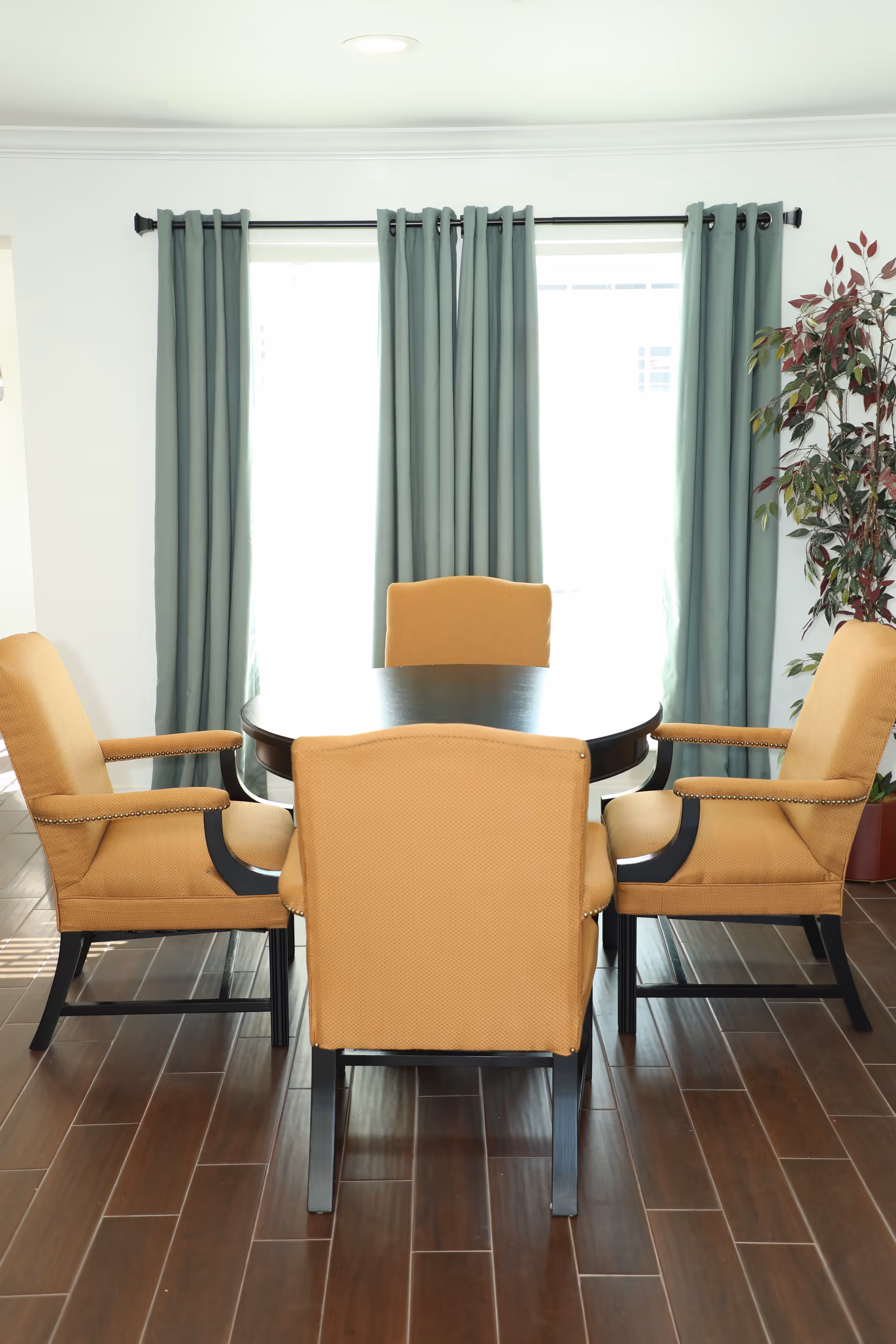 Round dark dining table surrounded by four mustard-colored upholstered chairs in front of a large window with teal curtains and a potted plant.