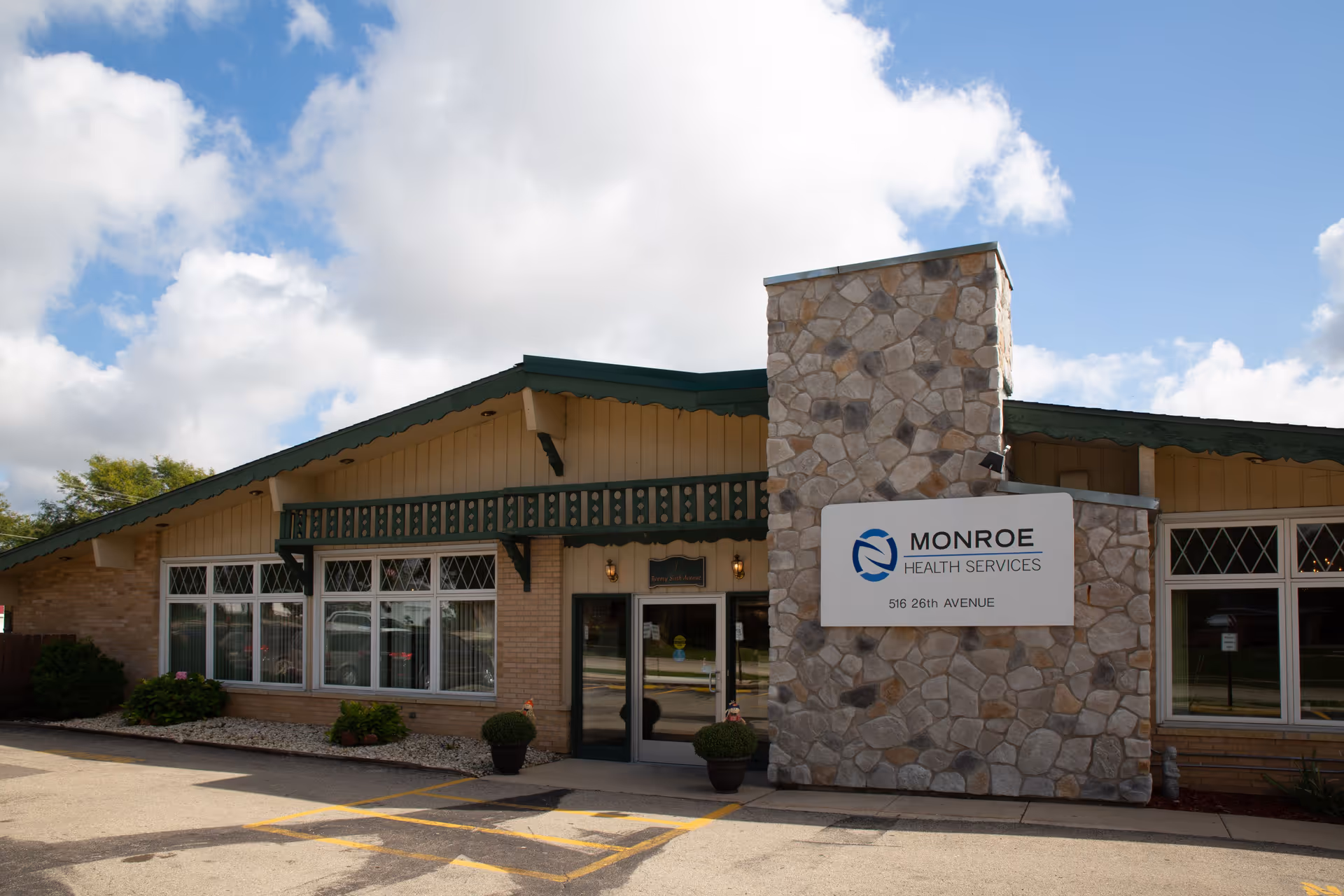 Front entrance of Monroe Health Services building with a stone facade sign, large windows, and a partly cloudy sky overhead.