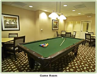 A game room featuring a pool table with a cue stick and billiard balls arranged on the green felt surface. Surrounding the pool table are several tables and chairs. The room has patterned carpet flooring, framed artwork on the walls, and ceiling lights.