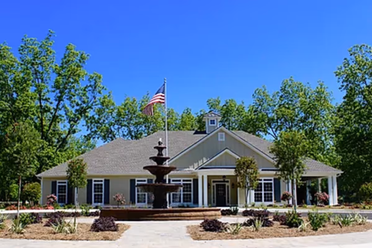 Front view of a single-story senior living building with a fountain and flag, surrounded by trees.