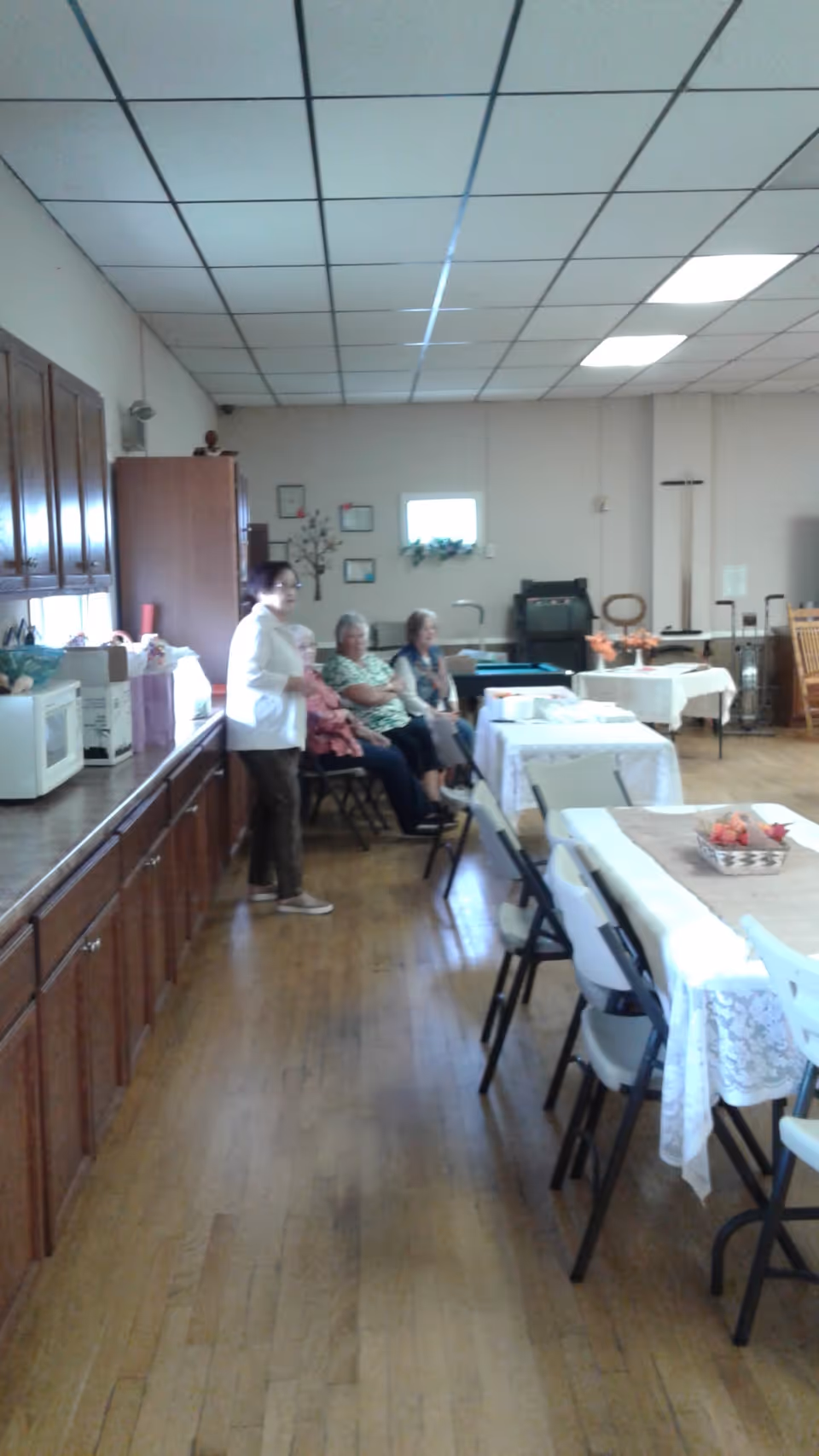 A staff member stands near seated residents in a communal dining room with kitchenette.