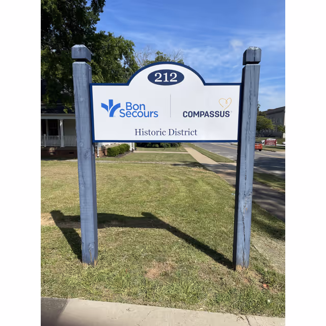 Blue and white sign on posts in a grassy area, showing Bon Secours and Compassus logos.