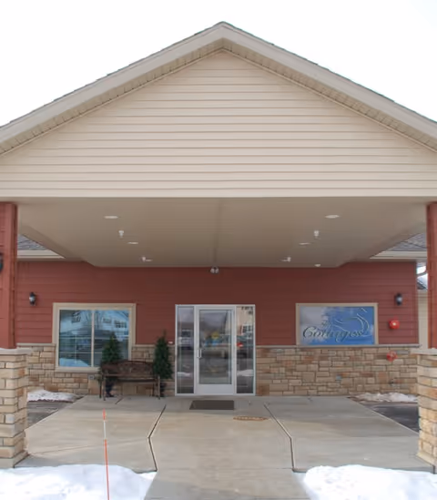Entrance of The Cottages on Golden Pond Memory Care facility with a covered driveway, stone and brick exterior walls, a glass door entrance, a bench, and a sign displaying the facility's name on the right side.