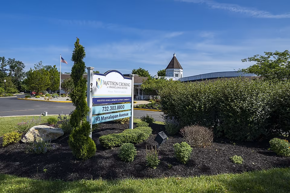Entrance sign for Mattison Crossing at Manalapan Avenue surrounded by landscaping with the facility building and an American flag in the background.
