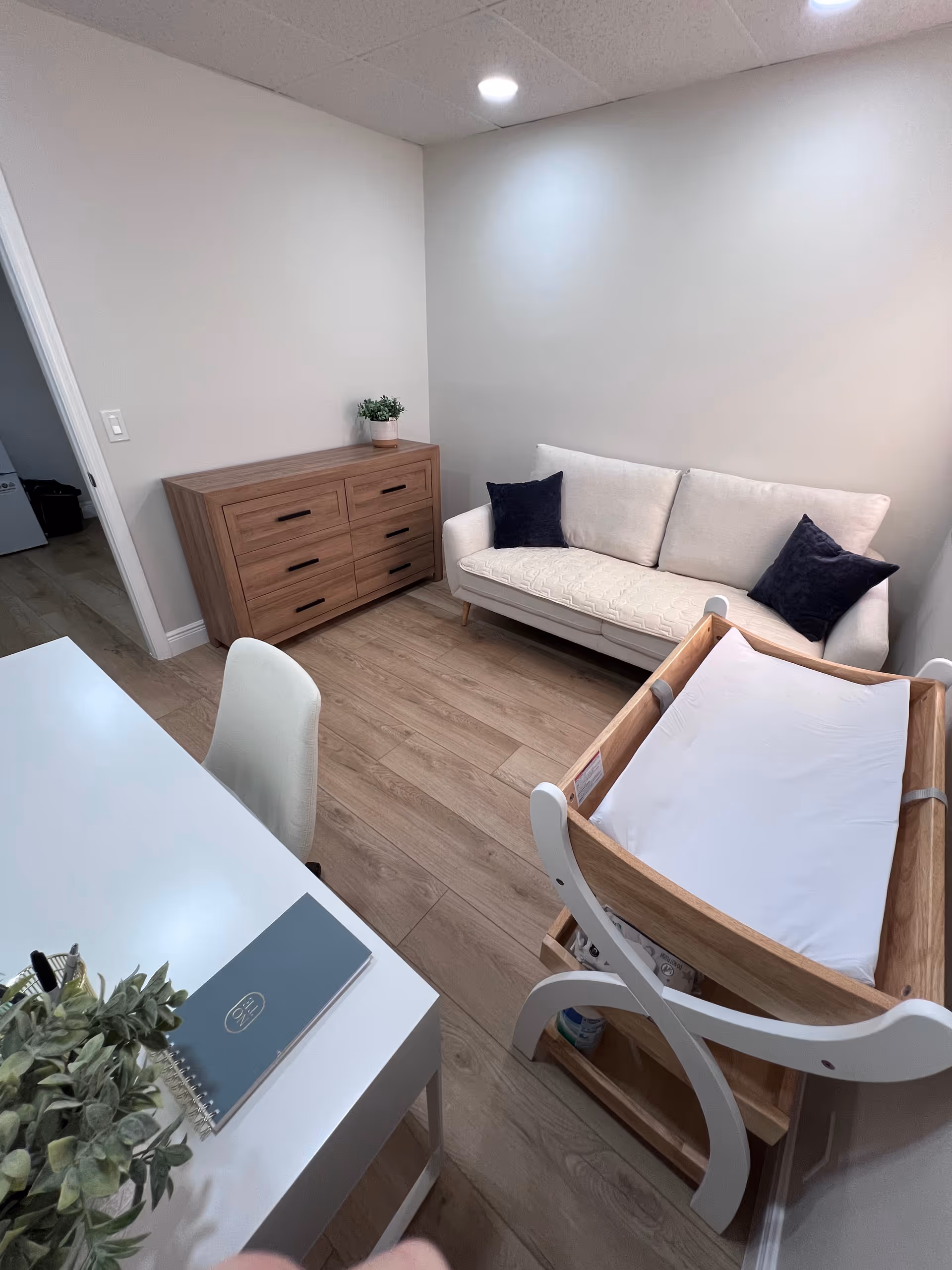 A small, modern interior with a white sofa, wooden dresser, and wooden baby cradle.