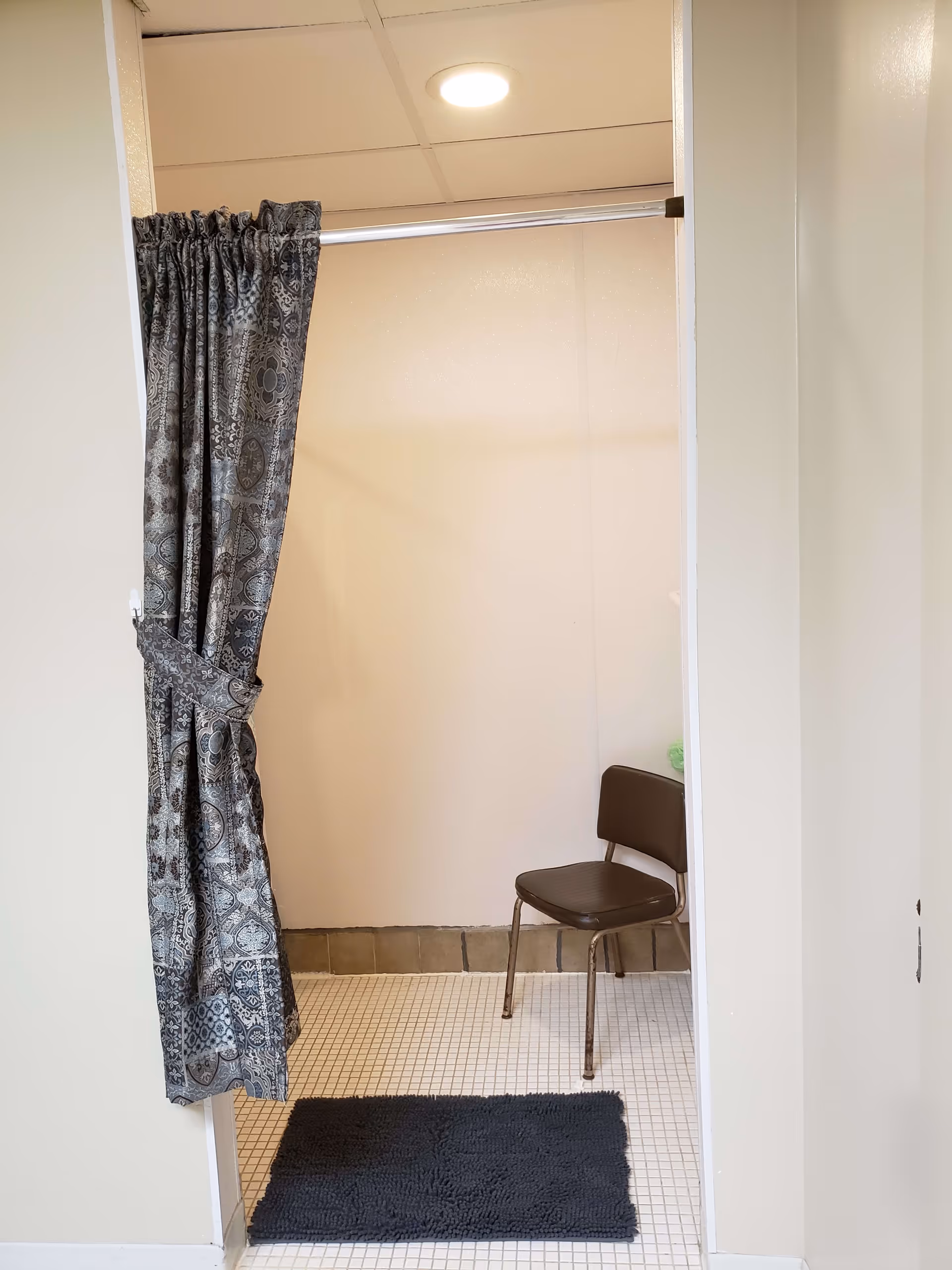 Small tiled bathroom/changing area with a patterned curtain tied back, a brown chair, and a black bath mat.