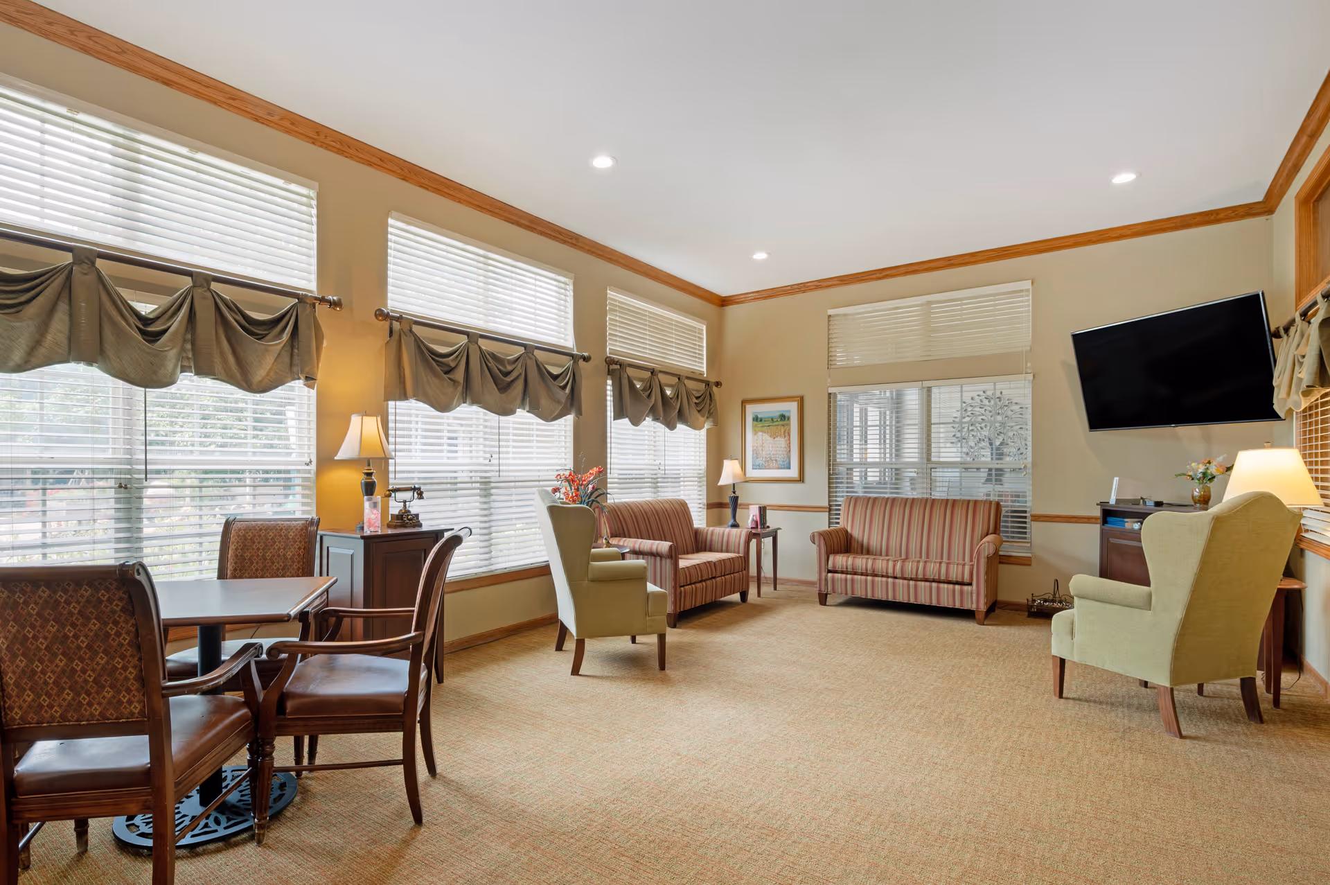 Bright senior living common area with seating, windows, lamps, and a wall-mounted TV.