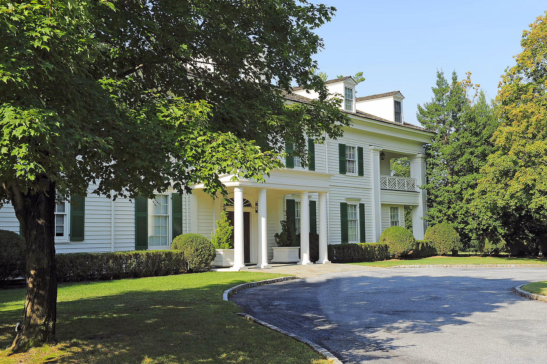 White two-story senior living facility exterior with columns, driveway, and green trees.