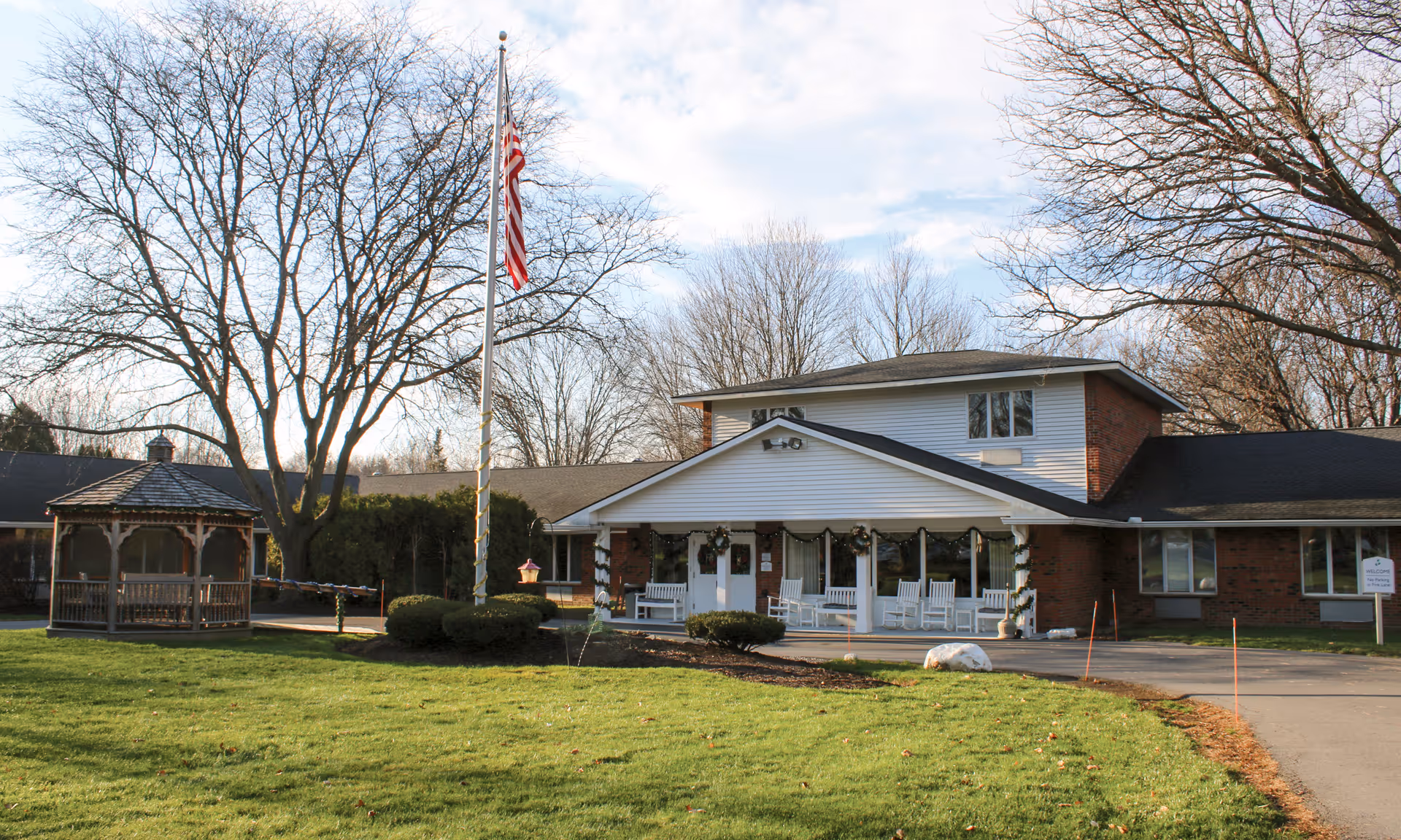 Front exterior of Brookdale Liverpool showing flagpole, white porch with rocking chairs, and gazebo.