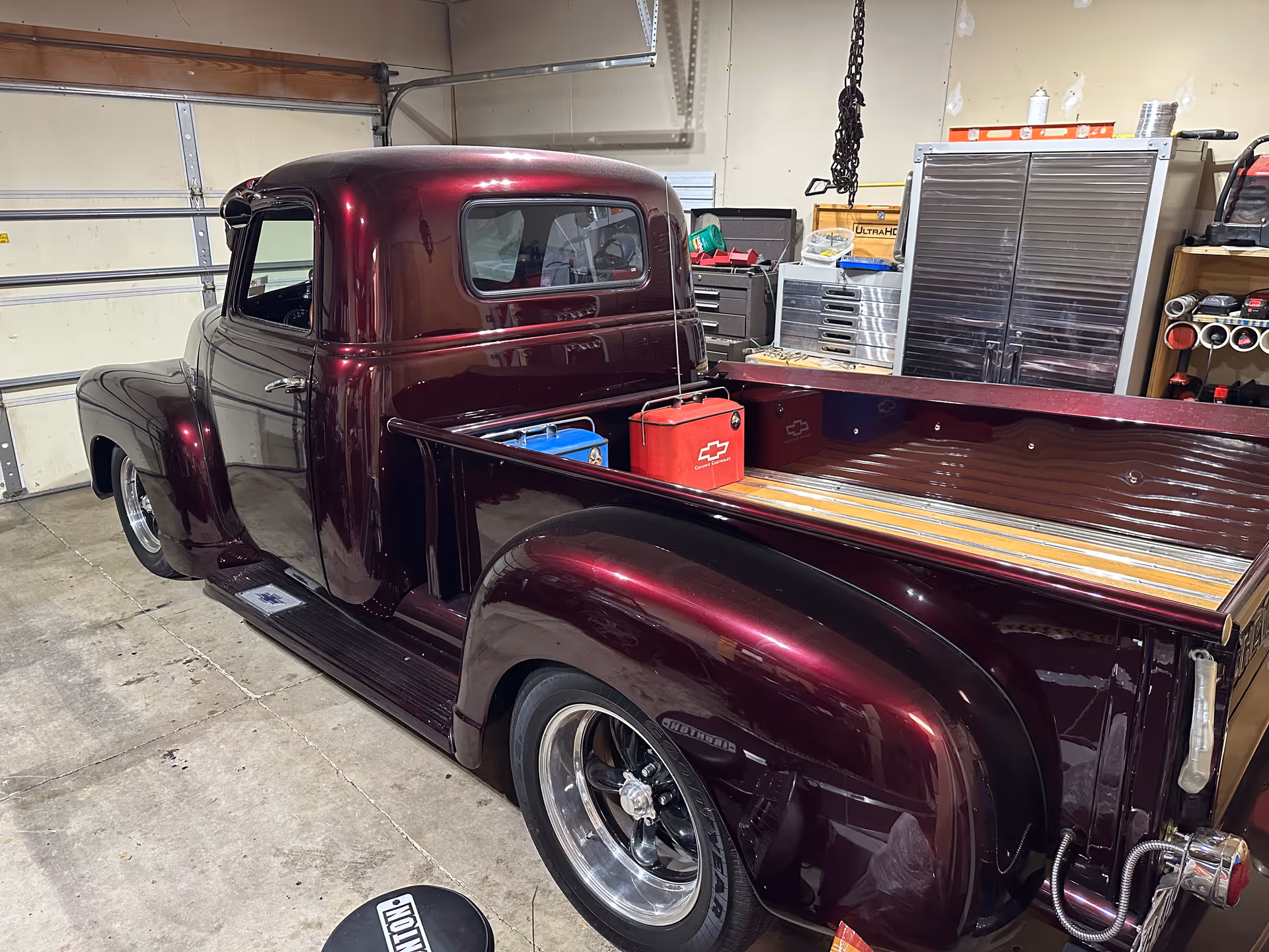 A vintage maroon pickup truck sits inside a cluttered garage with tools and cabinets.