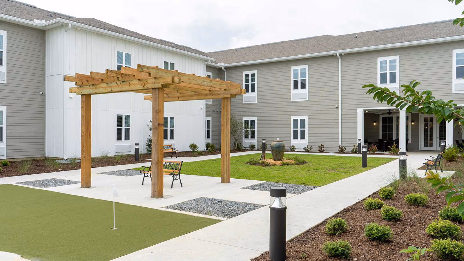 Outdoor courtyard area of a senior living facility featuring a wooden pergola with benches underneath, a small putting green with a golf hole flag, landscaped garden beds with shrubs, a central water fountain, and a two-story building with multiple windows surrounding the courtyard.