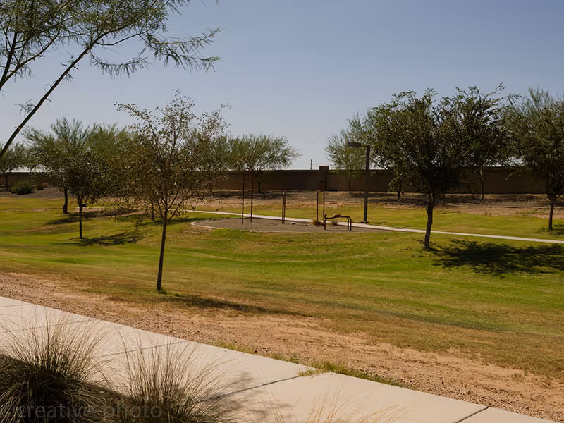 Outdoor area with green grass, several trees, a paved walkway, and exercise equipment under a clear blue sky.