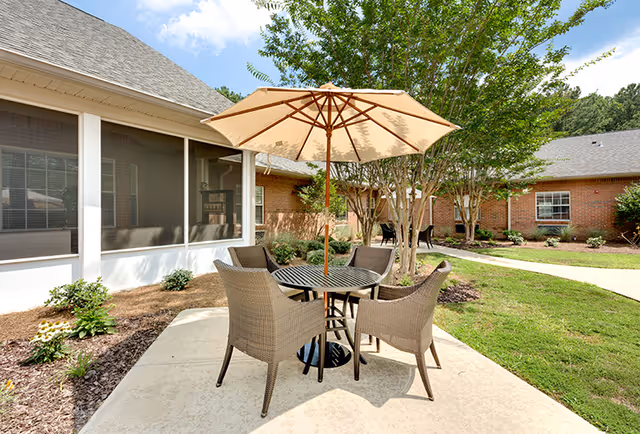 Outdoor courtyard patio with a round table, four wicker chairs and a large umbrella beside brick building wings and landscaping.
