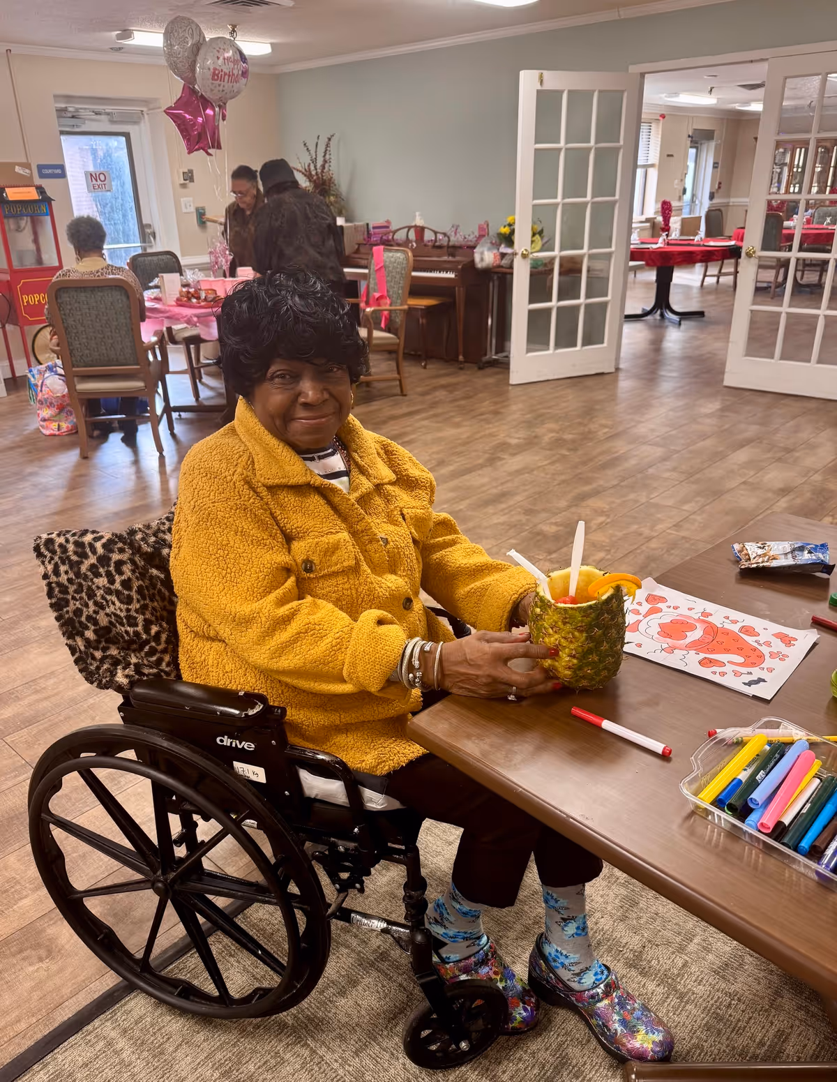 An elderly woman in a wheelchair wearing a yellow jacket and colorful shoes sits at a table in a communal room. She is holding a pineapple cup with a straw and has coloring markers and a coloring sheet on the table. In the background, other elderly people are seated at tables decorated with pink balloons and birthday decorations. The room has wooden flooring, a piano, and glass-paneled double doors leading to another room with more tables.