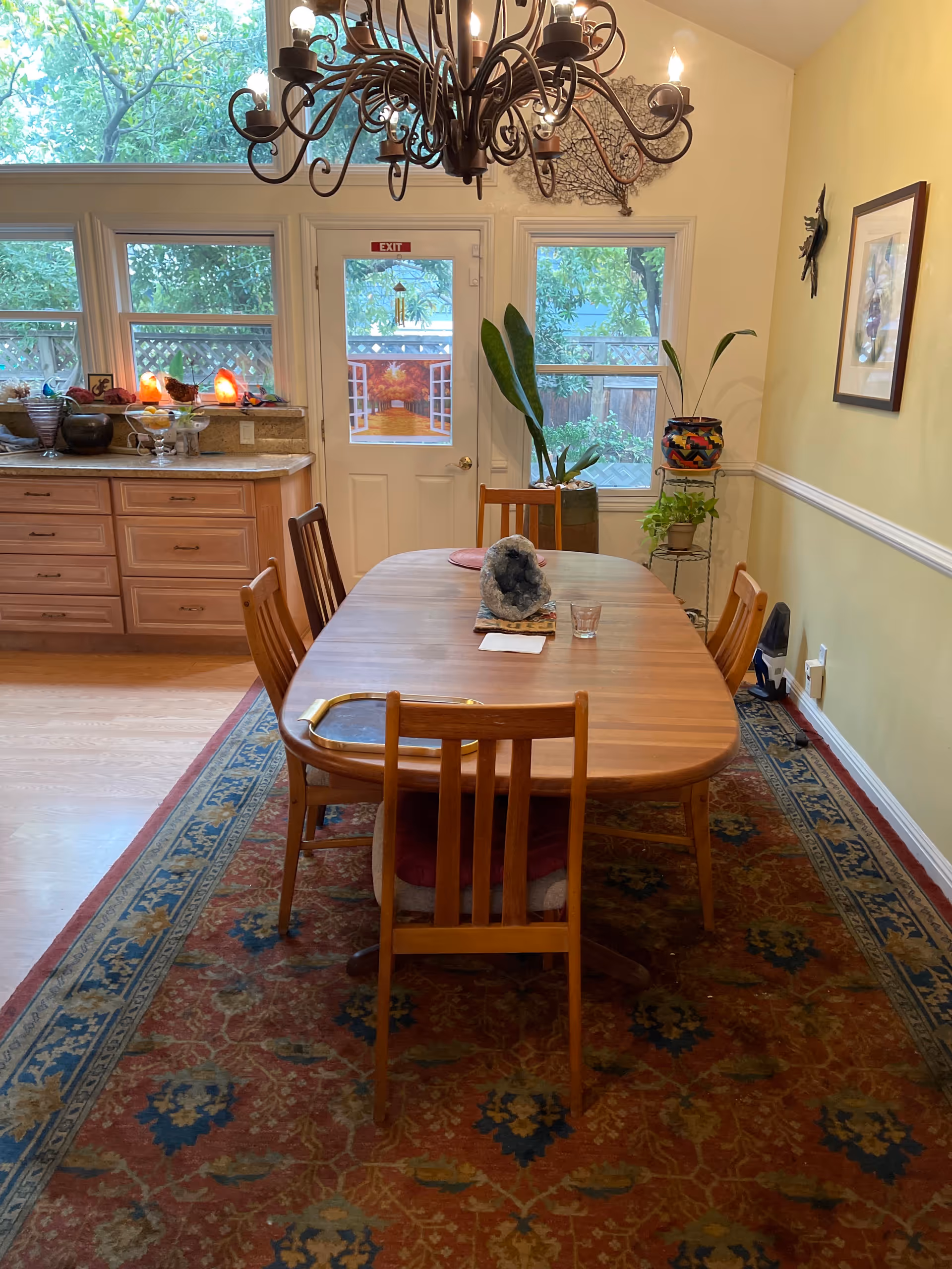 Wood dining table and chairs in a cozy dining room with chandelier, windows, and plants.