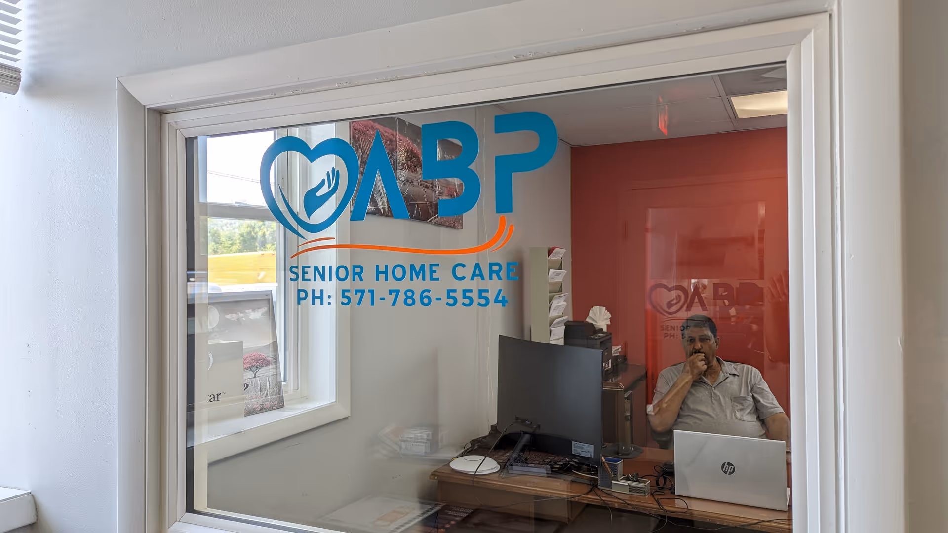 Office reception area visible through glass with ABP Senior Home Care logo and man at desk.