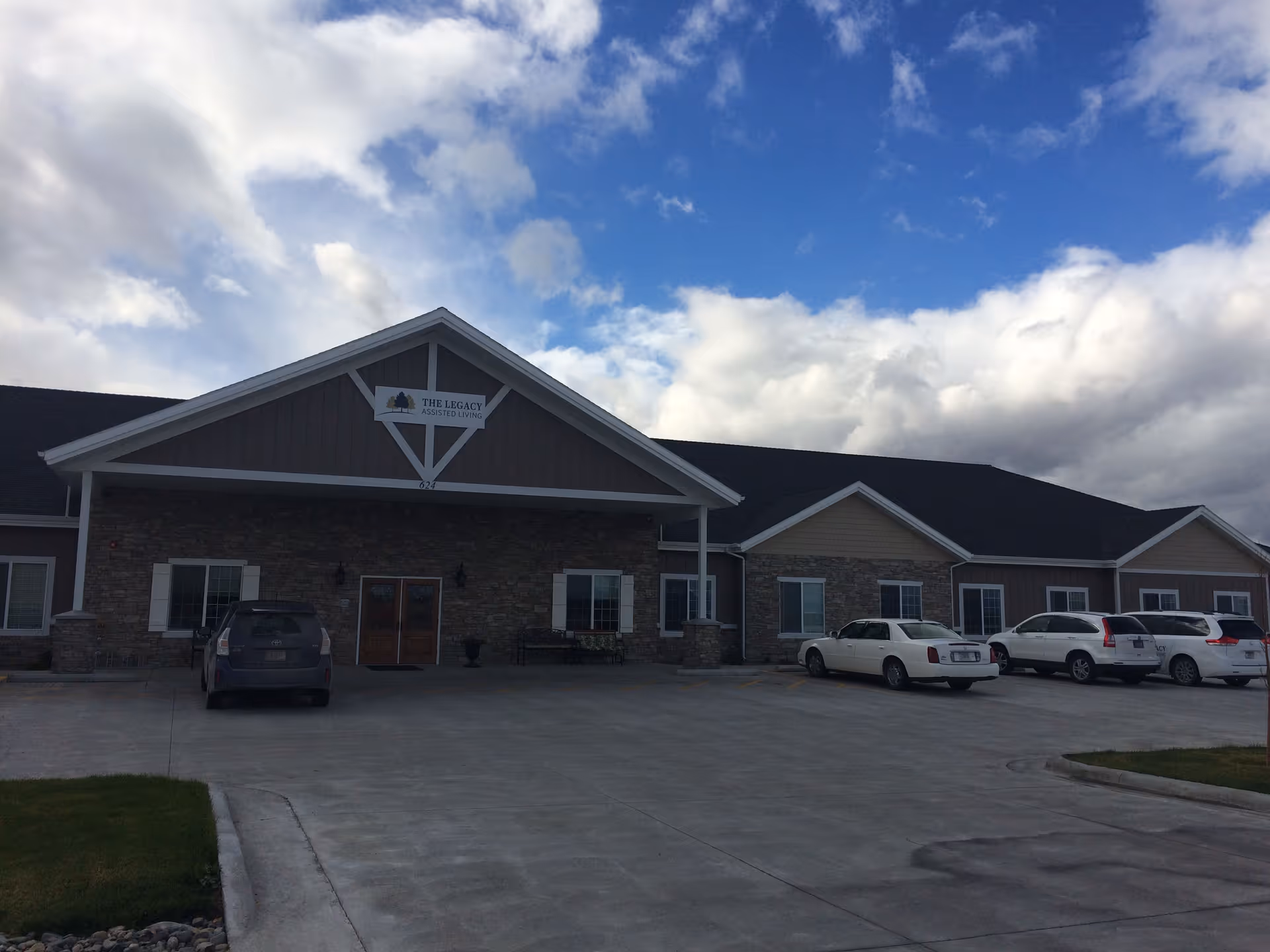 Exterior front view of The Legacy Assisted Living facility showing a single-story building with stone and siding facade, multiple windows, a covered entrance, and several parked cars in the driveway under a partly cloudy sky.