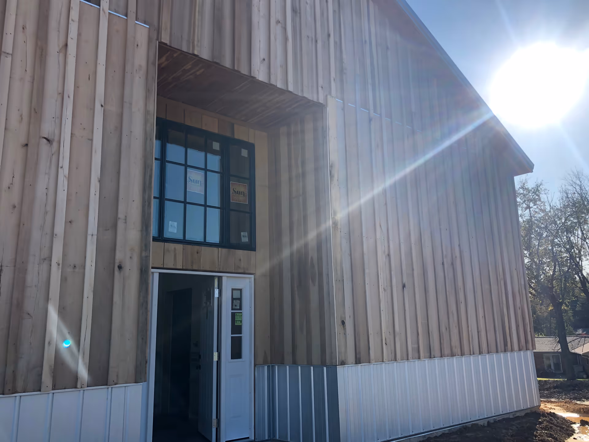 Exterior view of a building under construction with vertical wooden siding and a white door partially open. There is a large window above the door with stickers on the glass. The sun is shining brightly in the sky, creating lens flare in the image.