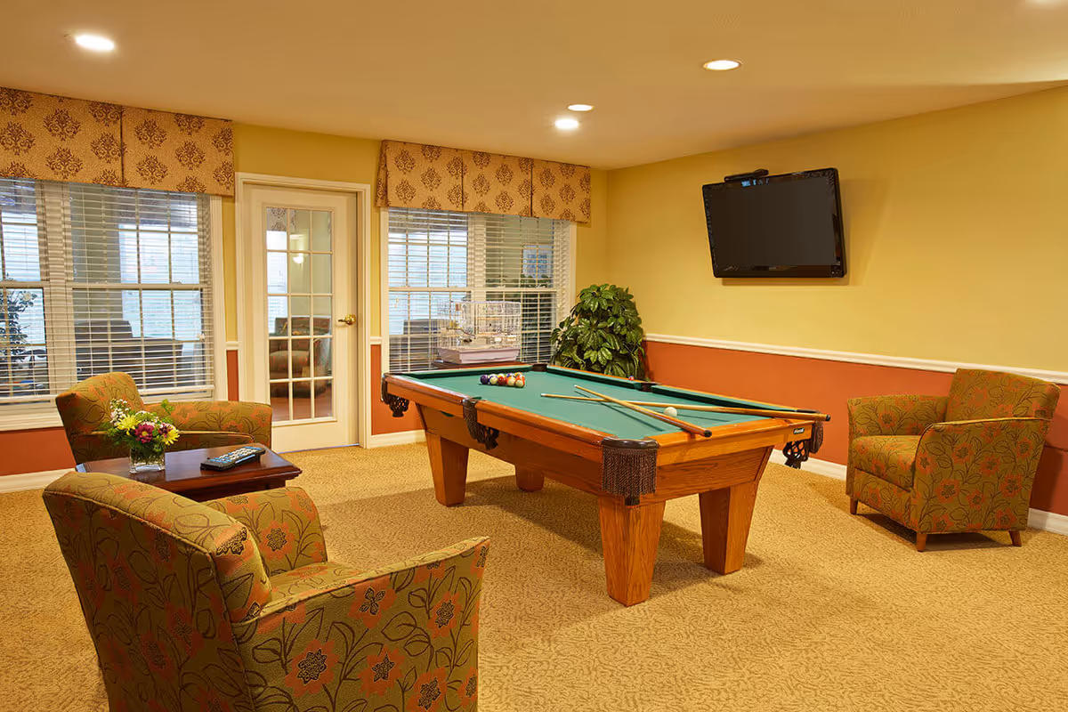 A cozy interior game room with a pool table in the center, upholstered chairs around a small table, a wall-mounted TV, and windows with blinds.