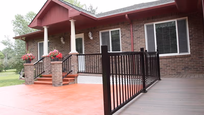 Front exterior view of a brick building with a covered porch supported by white columns, red steps leading up to a white door, black metal railings, and flower pots with pink flowers on brick pillars. There is a spacious red concrete patio and a wooden deck area with a black railing. Green trees and grass are visible in the background.
