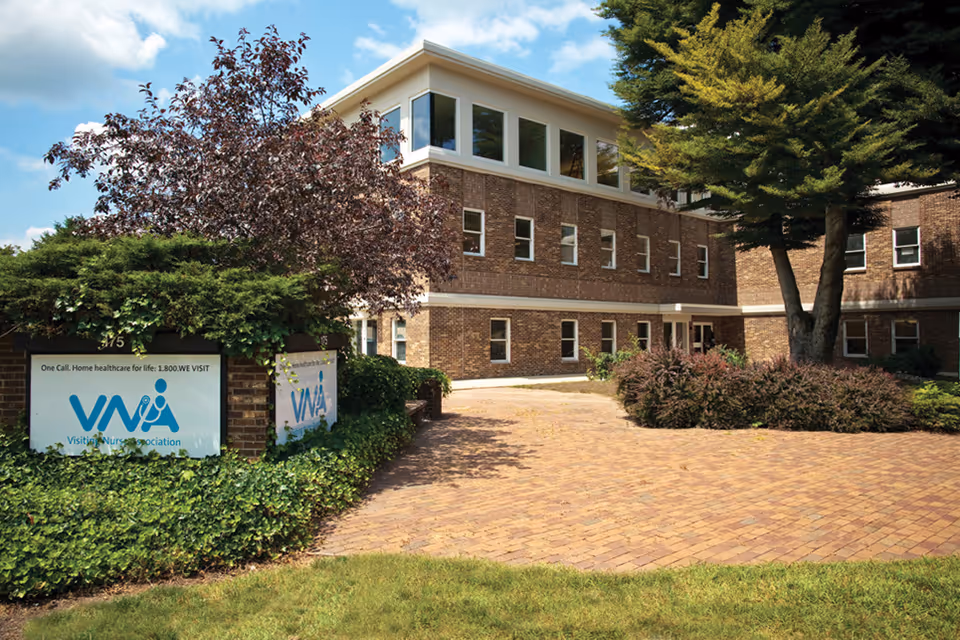 Brick nursing facility building with a paved entrance, trees, and a visible VNA sign.