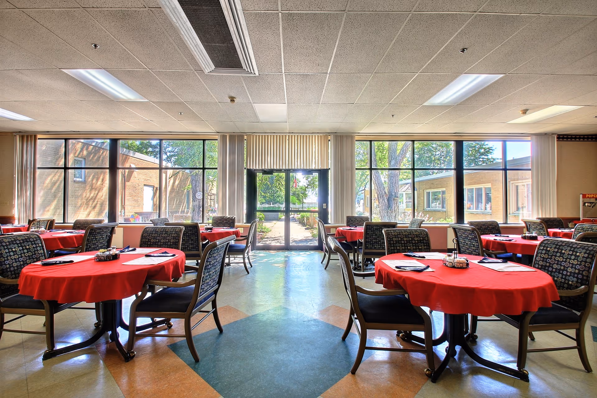 A bright dining room with several round tables covered in red tablecloths, each surrounded by patterned cushioned chairs. Large windows and a glass door let in natural light and offer a view of the outdoor area with trees and buildings.