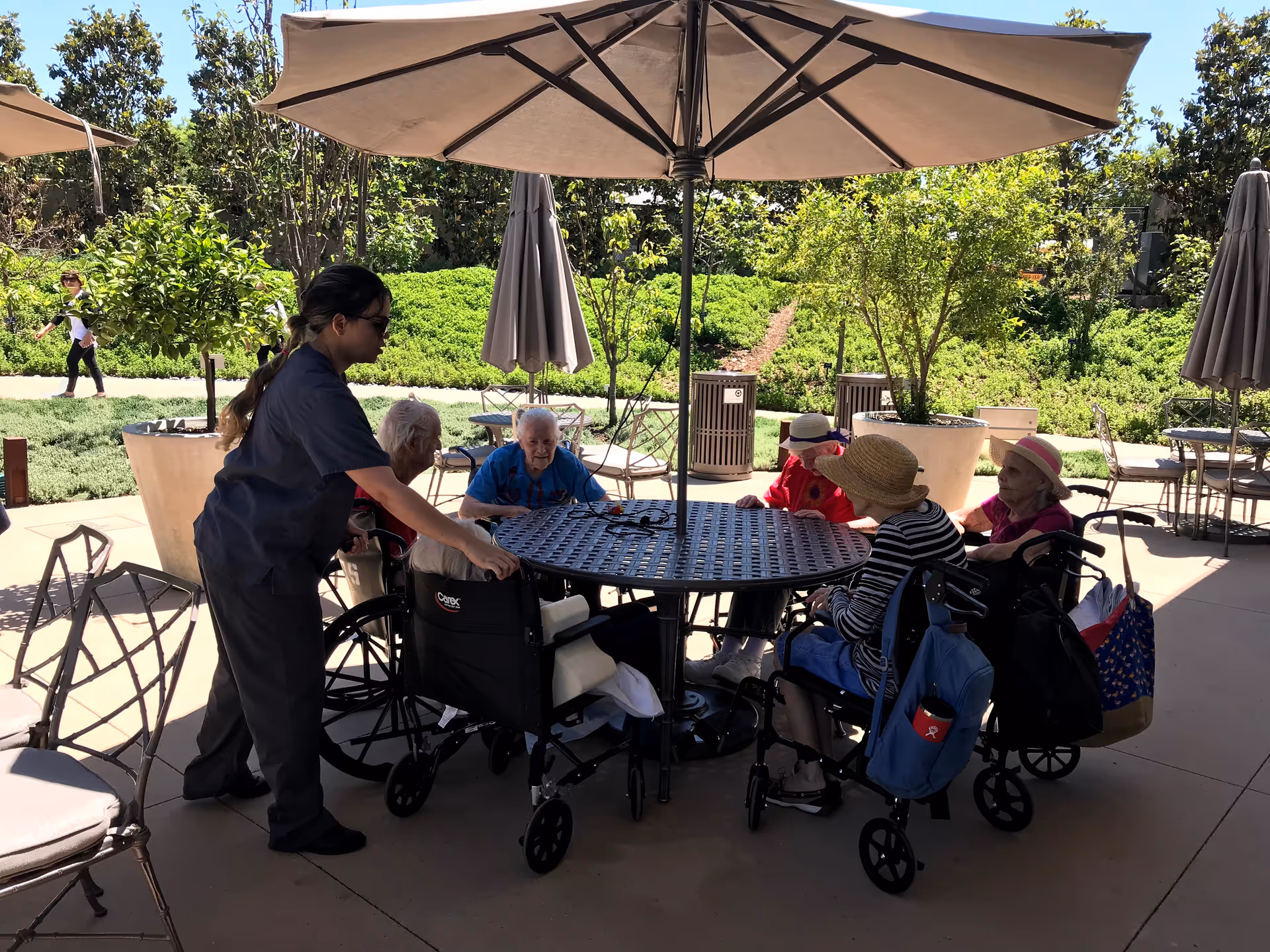 A caregiver assists elderly individuals seated around a round outdoor table under a large umbrella. The elderly people, some in wheelchairs, are engaged in conversation or resting. The setting is a sunny patio area with greenery and trees in the background.