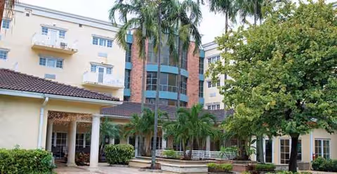 Exterior view of Emerald Park of Hollywood, a cream-colored senior living building with palm trees.