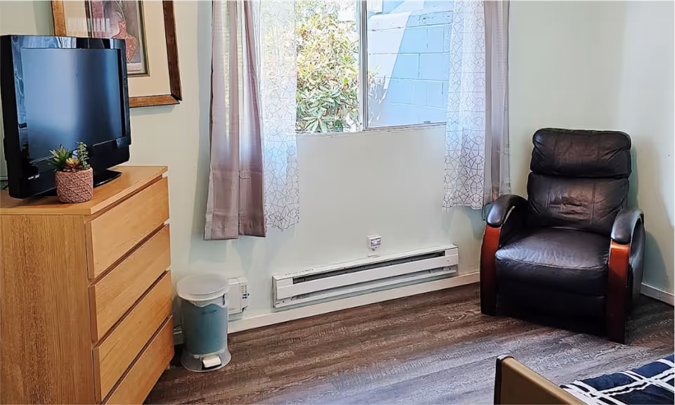 A small living room with TV on dresser, leather chair, window, and baseboard heater.