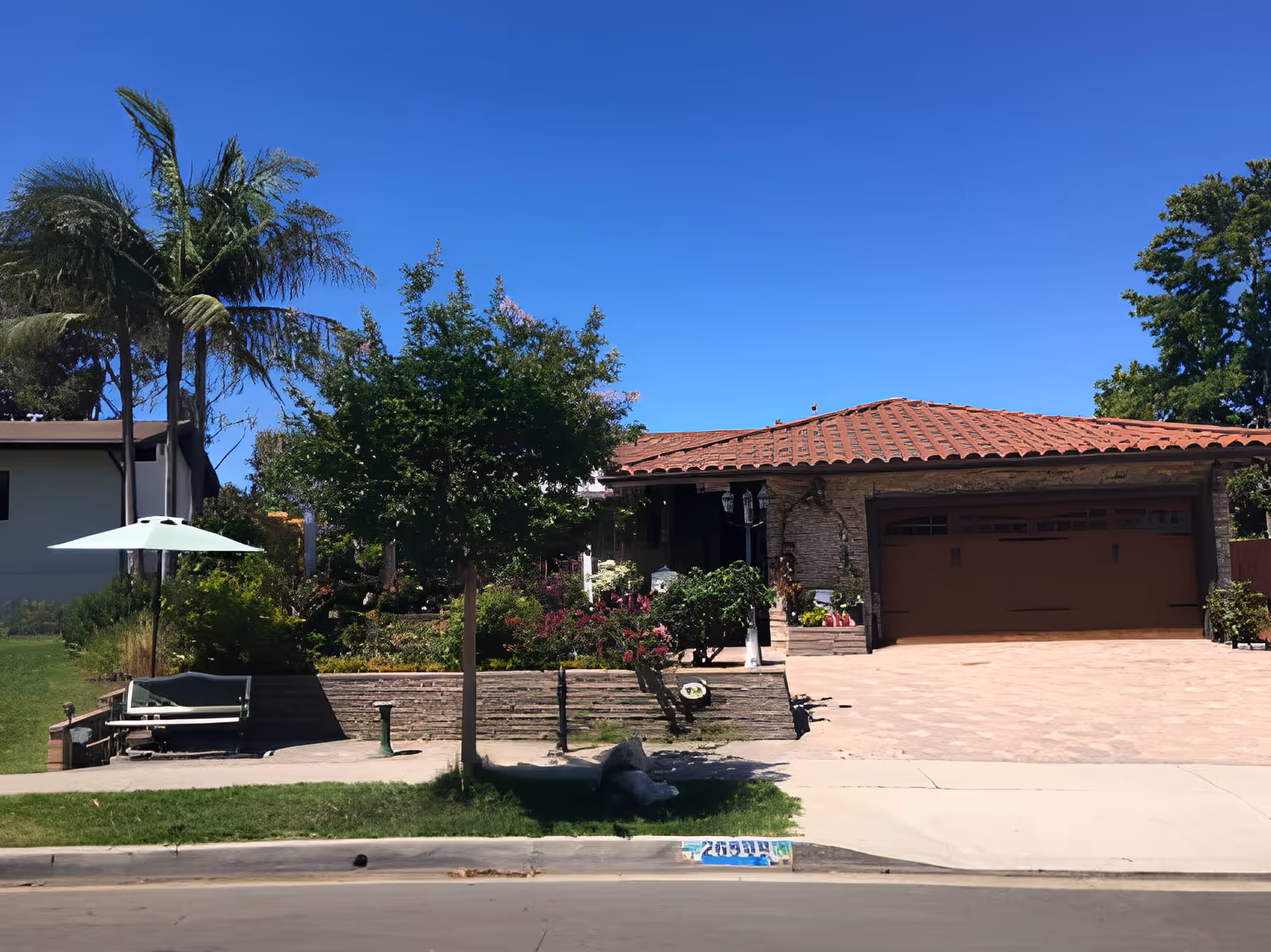 Exterior view of a single-story house with a red tiled roof, a large garage door, and a well-maintained garden with various trees and shrubs. There is a bench with a green umbrella on the left side near the sidewalk, and a clear blue sky overhead.