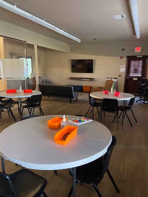 Open senior living common area with round tables, chairs, and a wall-mounted TV.