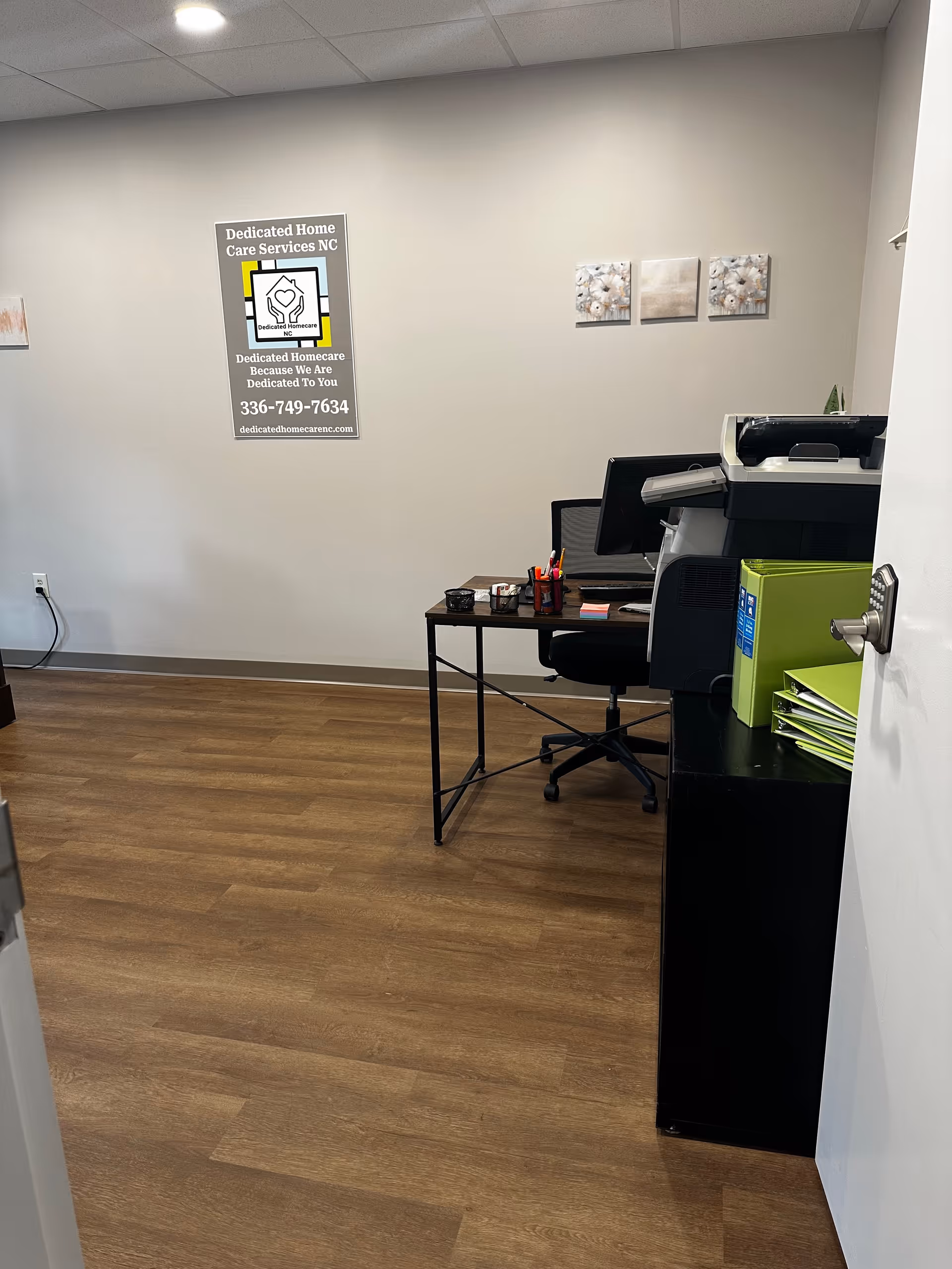 Office workspace with desk, computer, printer, and sign reading Dedicated Homecare NC.