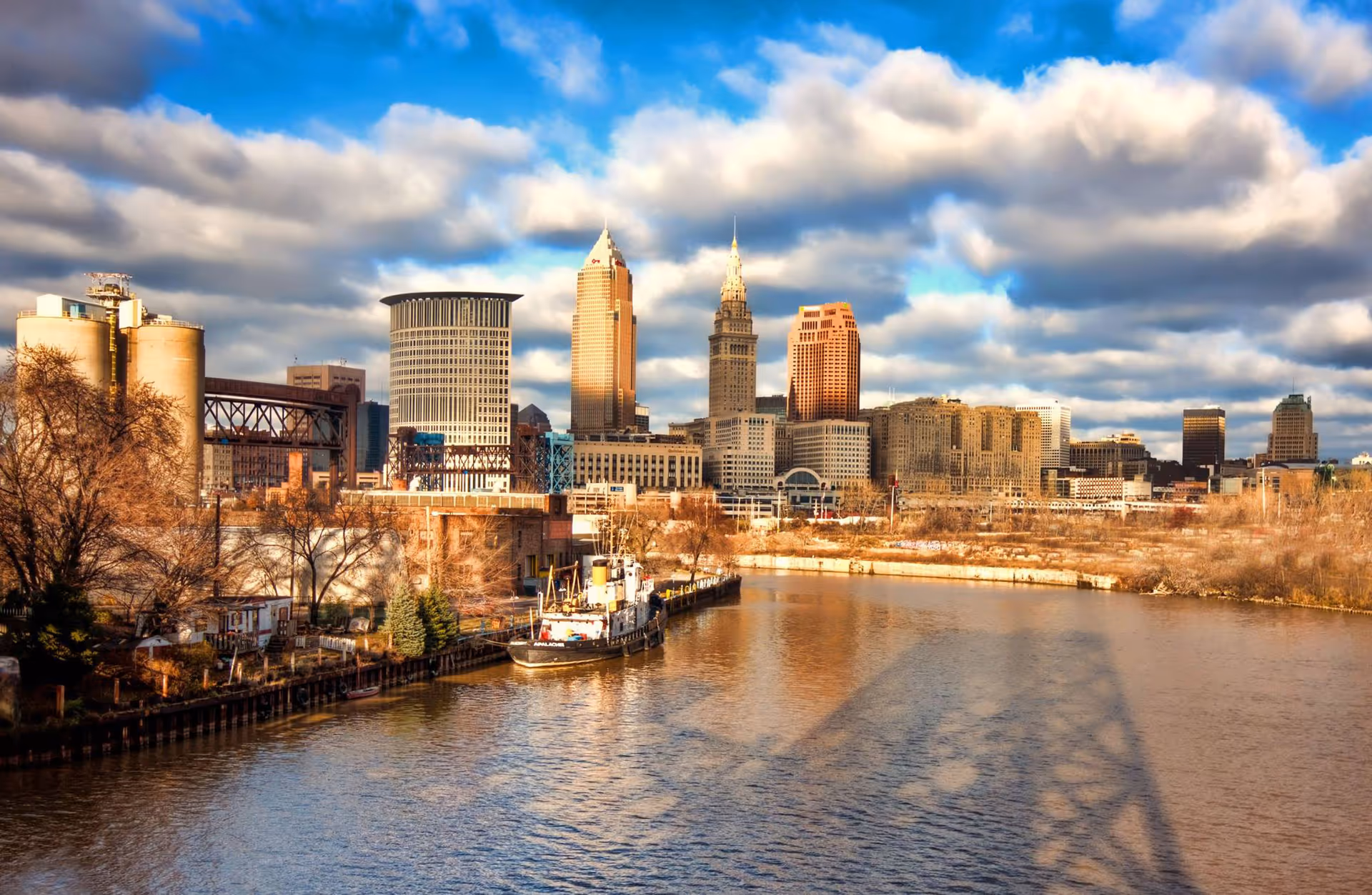 City skyline along a river with boats and a cloudy sky.