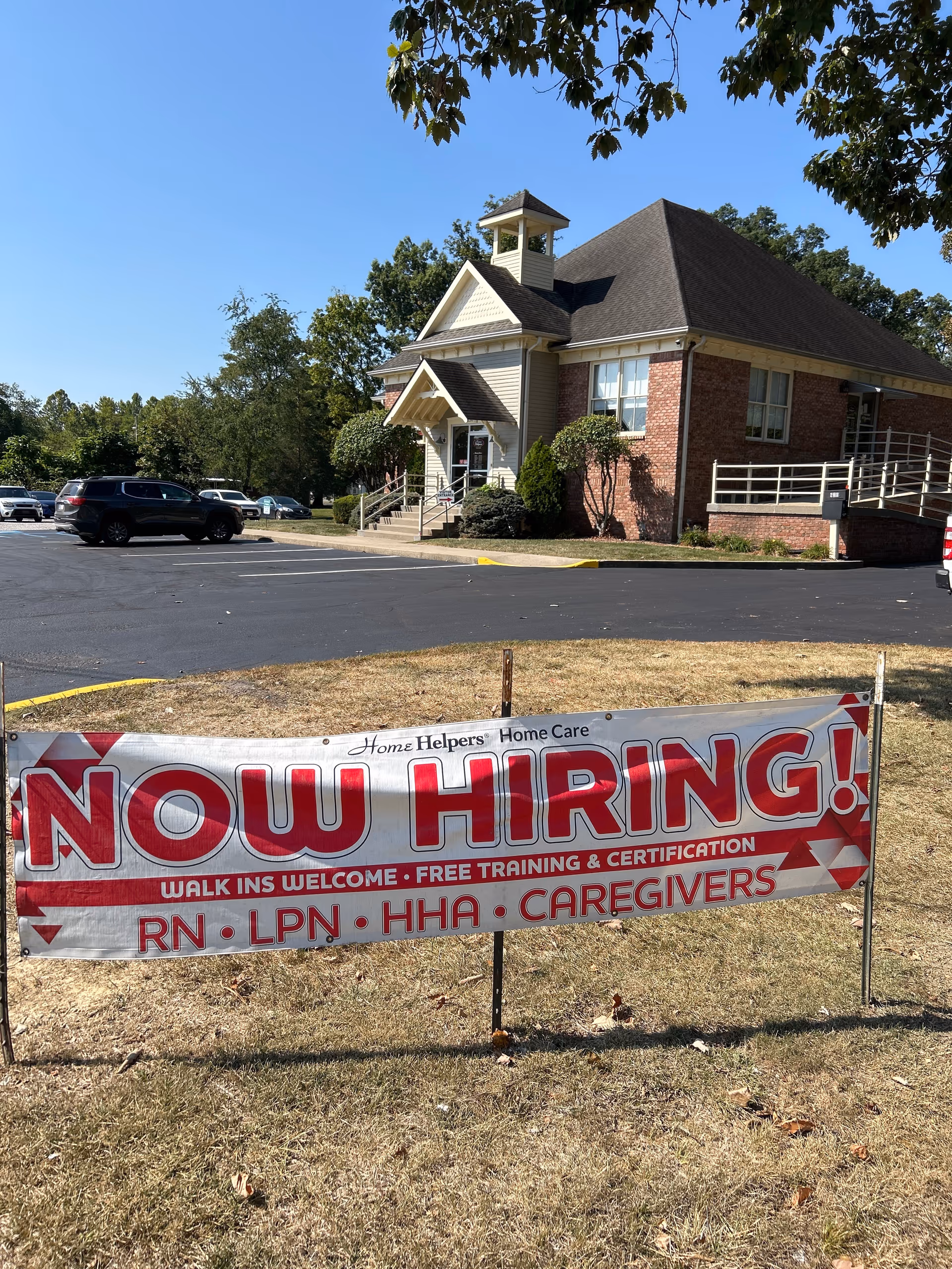 Exterior view of a brick senior care building with a large 'Now Hiring' banner in front.