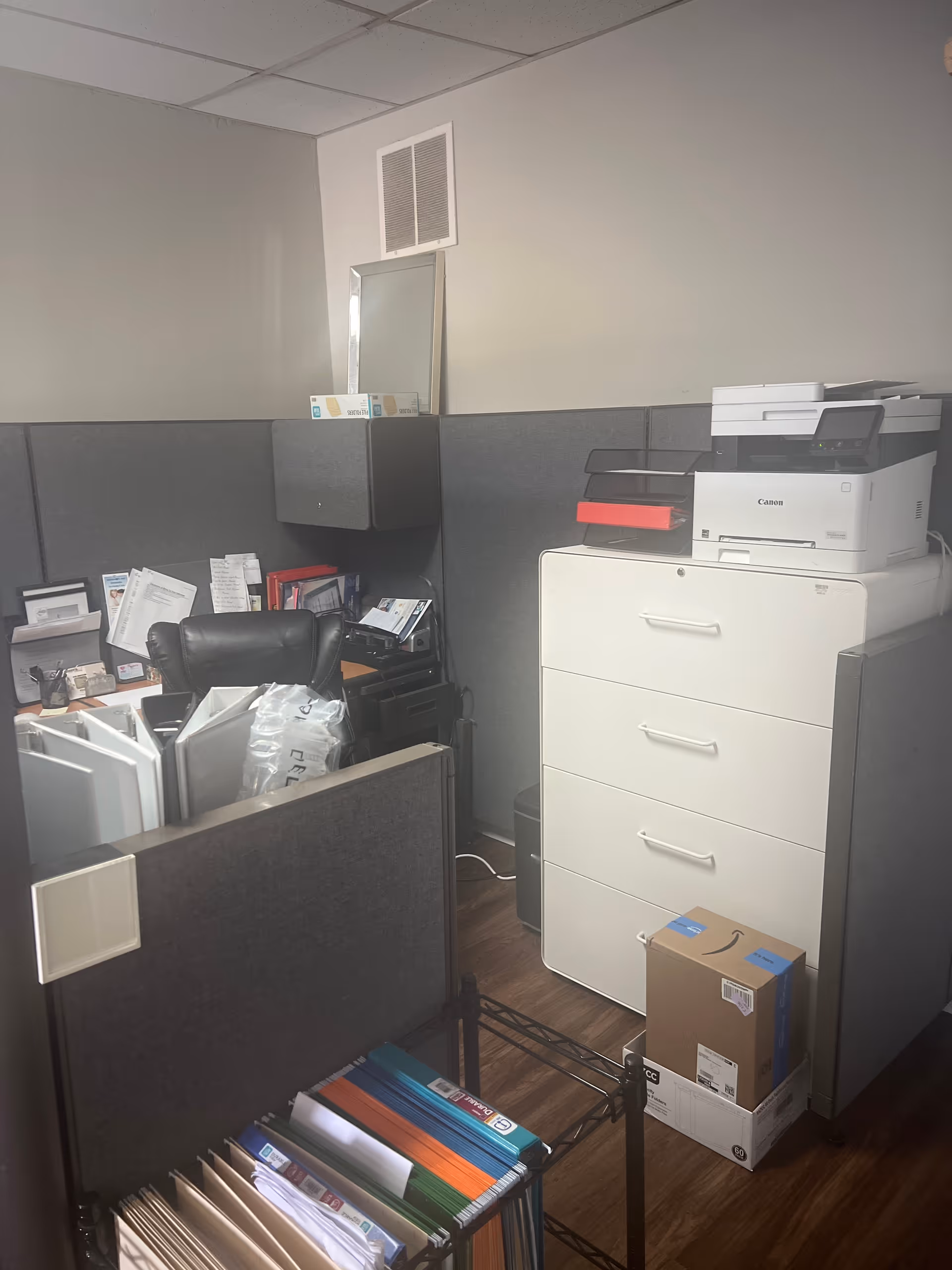 Office cubicle with a white filing cabinet, printer, and cluttered folders.