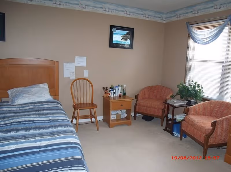 A bedroom with a single bed covered in a blue striped comforter, a wooden chair, a small wooden nightstand with various bottles on top, two upholstered armchairs, a small side table with a plant and magazines, and a window with blinds and a blue valance.