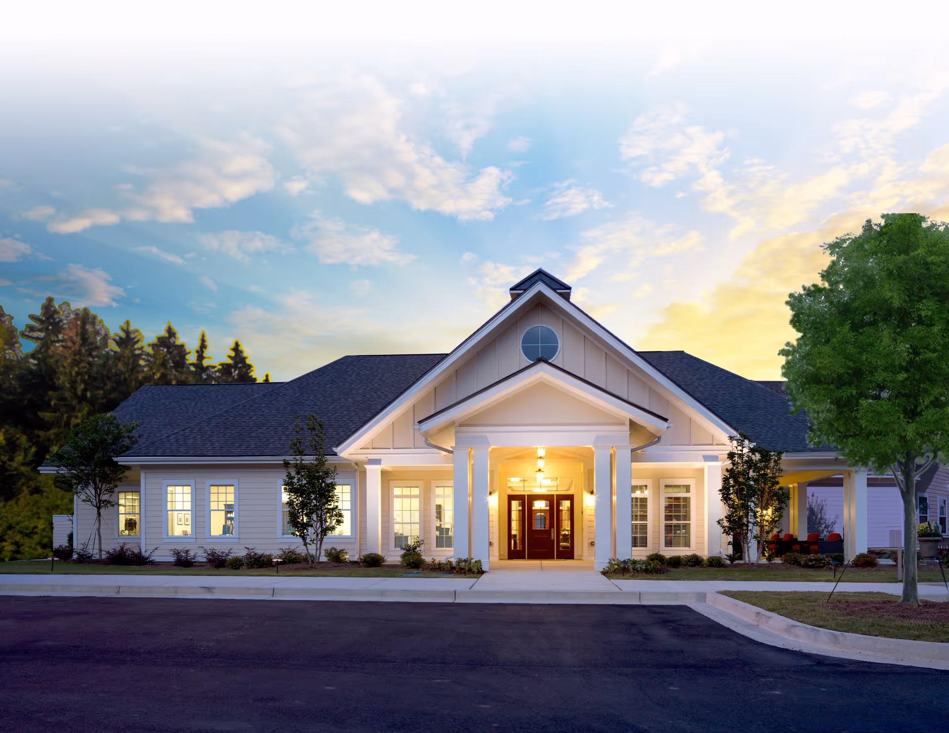Front exterior view of a single-story senior living community building with a peaked roof, white siding, large windows, and a covered entrance with columns. The building is surrounded by small trees and landscaping, with a paved driveway in front and a partly cloudy sky at sunset.