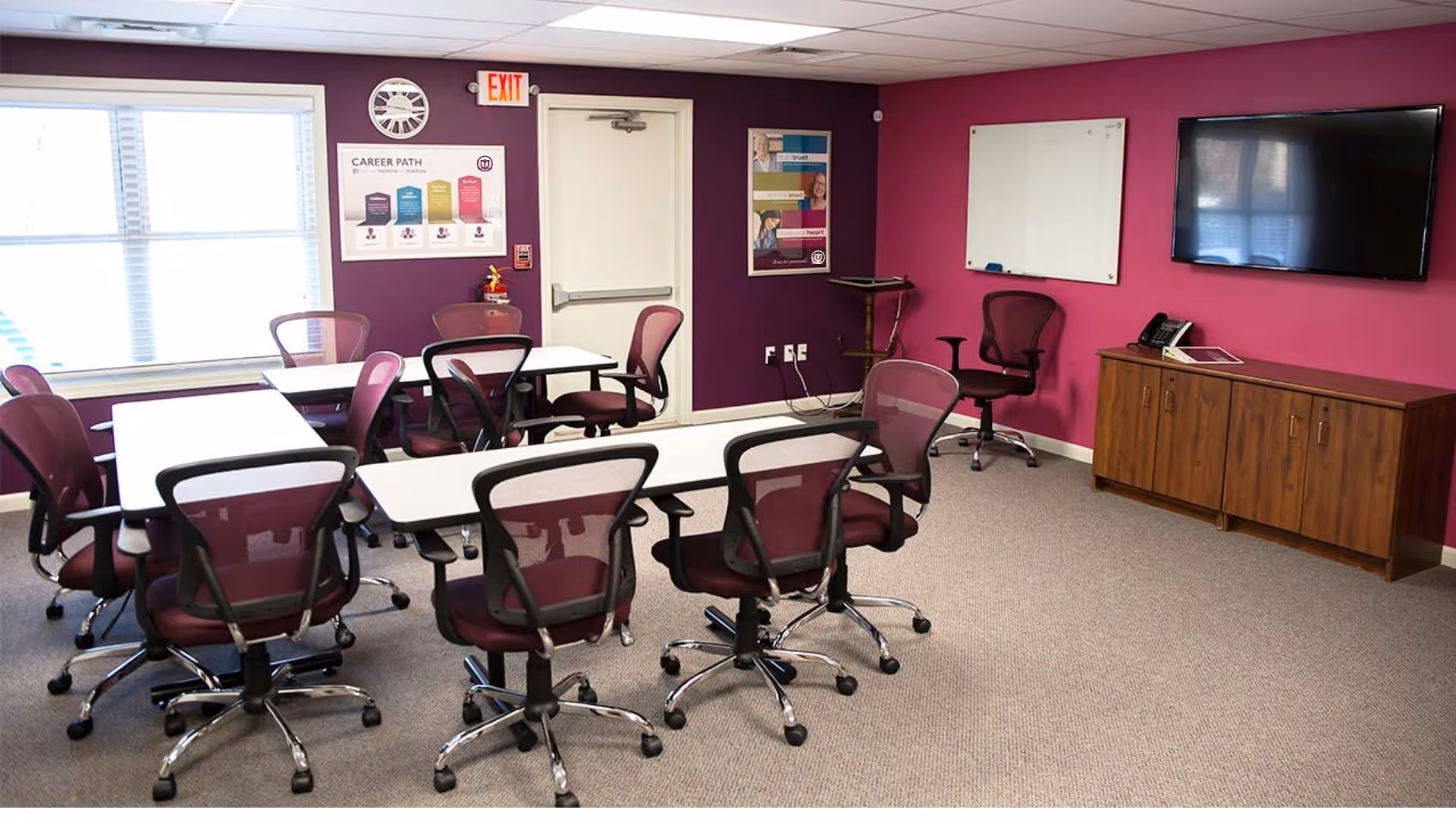 Magenta-walled conference room with tables, chairs, whiteboard, and a wall-mounted TV.