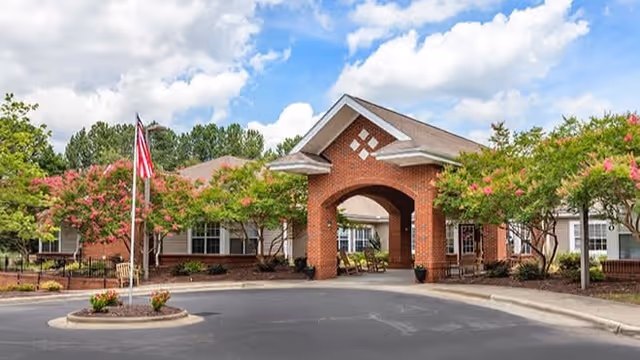 Front entrance of a brick senior living facility with a covered porte-cochere, American flag, and landscaped trees.