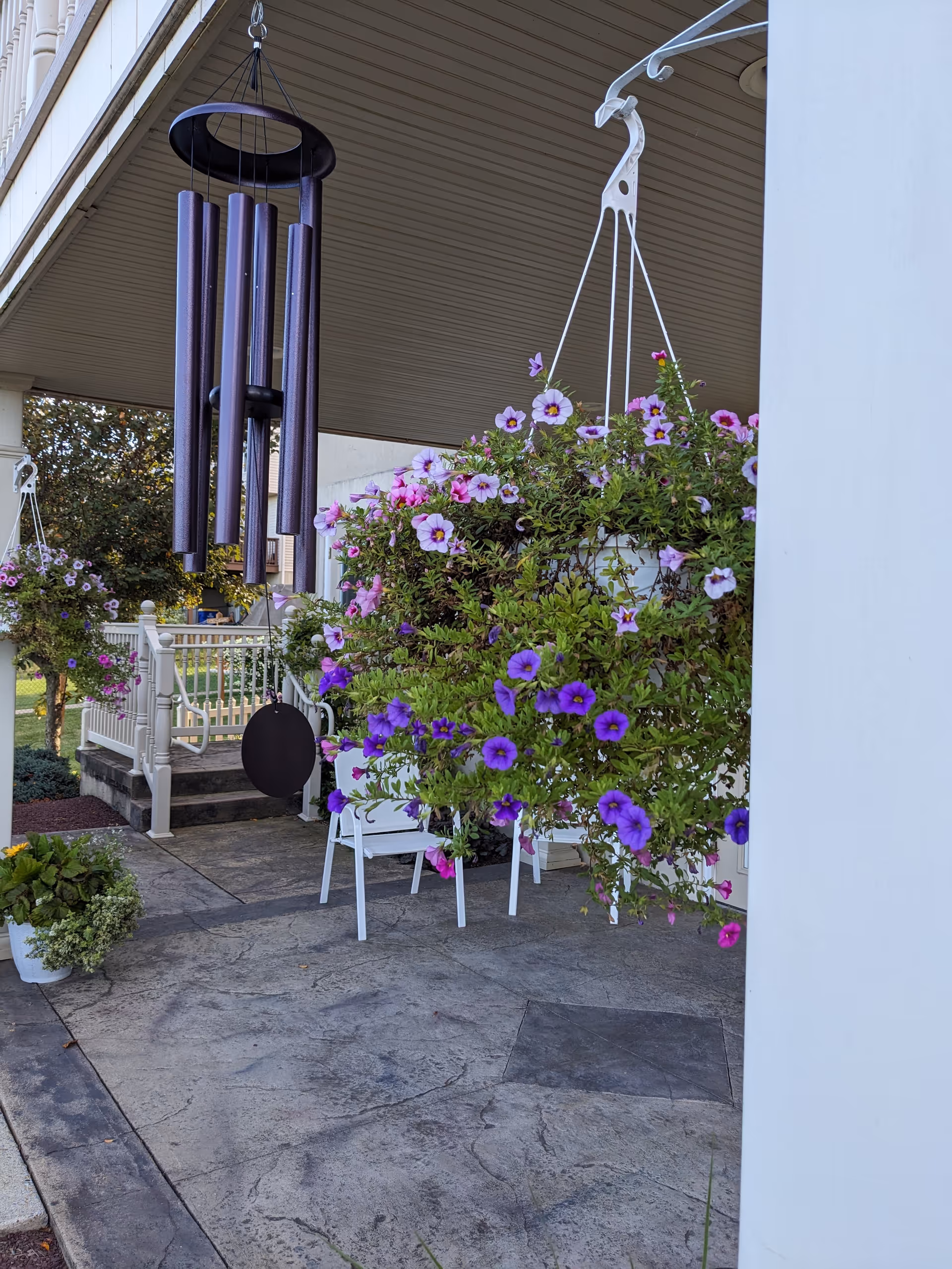 Hanging flower baskets, wind chime, and white chairs on a covered outdoor patio.