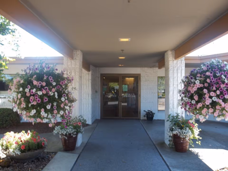 Entrance to Frontier Rehabilitation and Extended Care Center with double glass doors under a covered walkway. There are large hanging baskets filled with pink and white flowers on either side of the entrance, along with additional potted plants near the pillars.