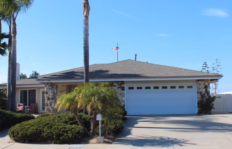 Front exterior of a single-story assisted living home with stone facade and palm trees.