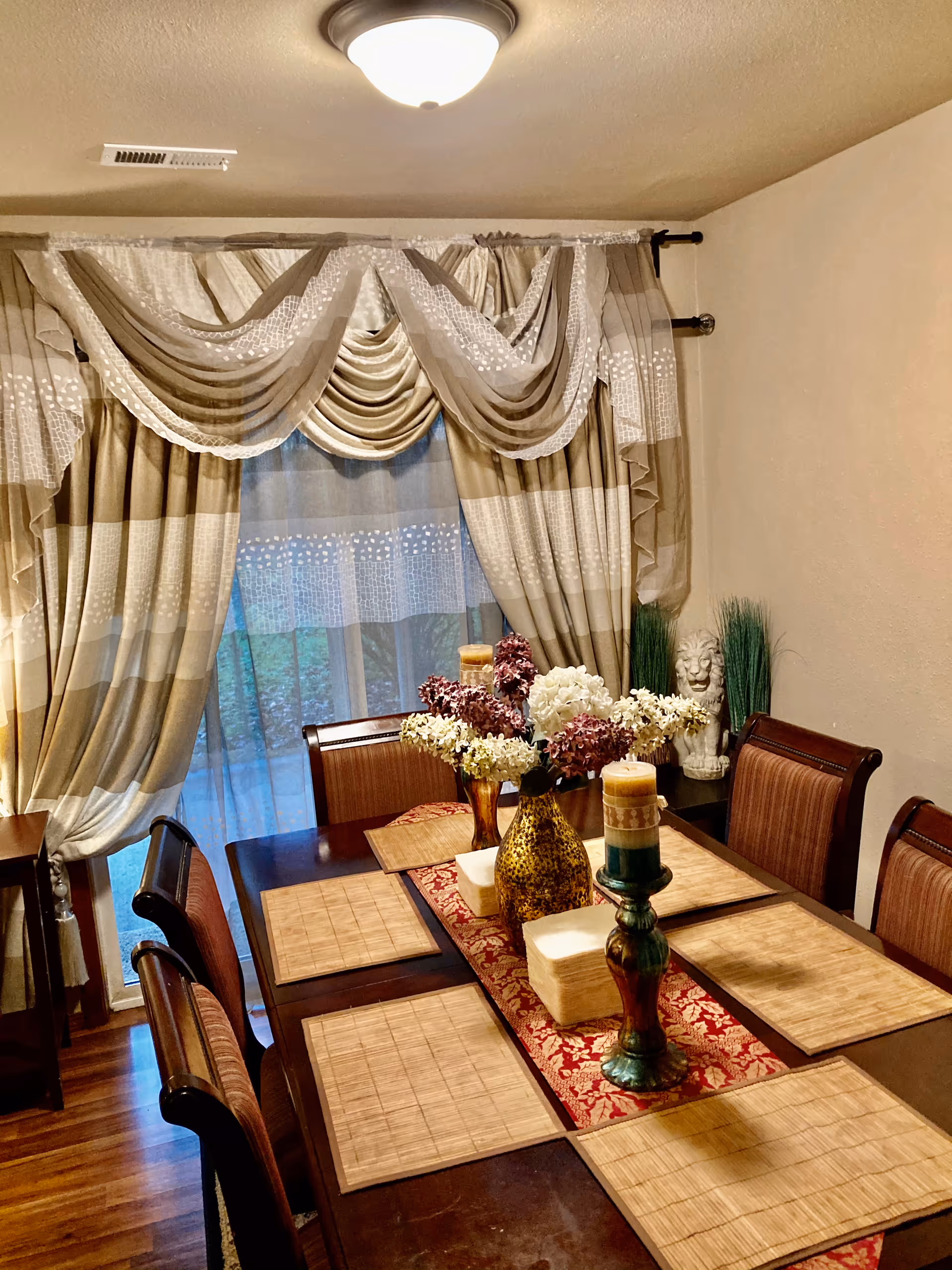 A dining room with a long table, chairs, floral centerpiece, and layered curtains.