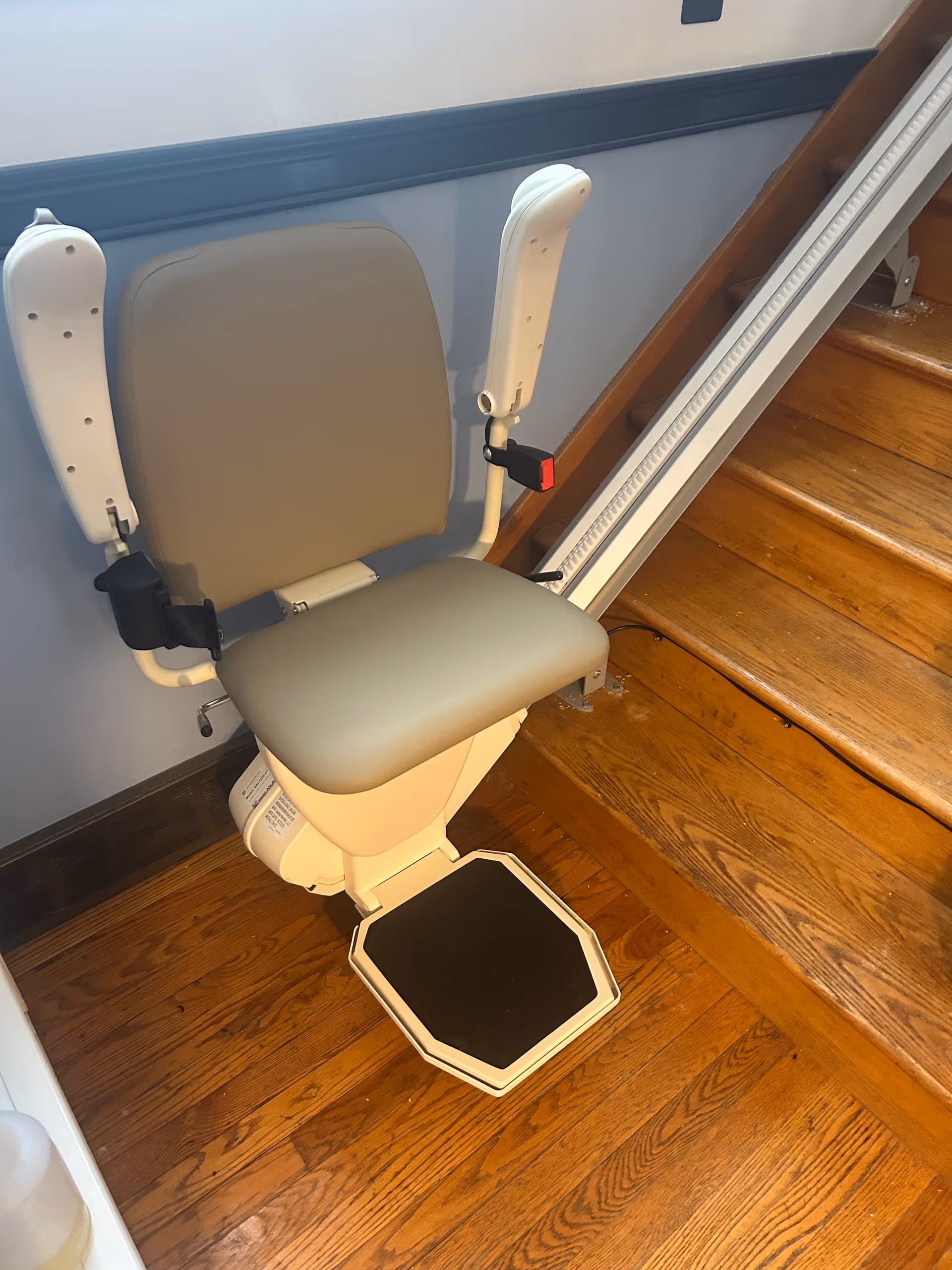 Indoors, a stairlift chair is mounted beside wooden stairs in a facility hallway.