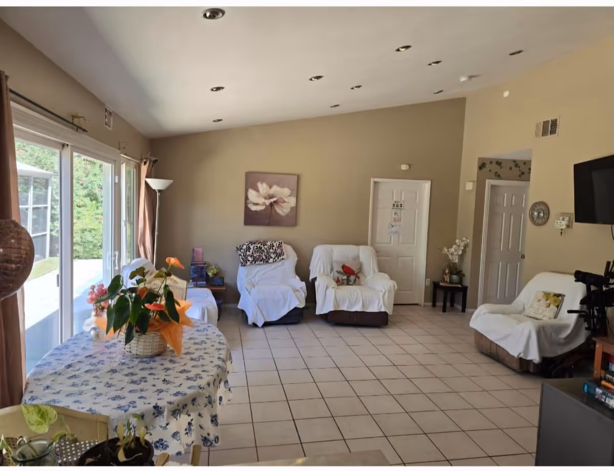 A bright living room with beige walls and tiled floor featuring three armchairs covered with white slipcovers, a round table with a floral tablecloth and potted plants, a floor lamp, a wall-mounted TV, and a door with a sign. Large sliding glass doors on the left open to an outdoor area with greenery.